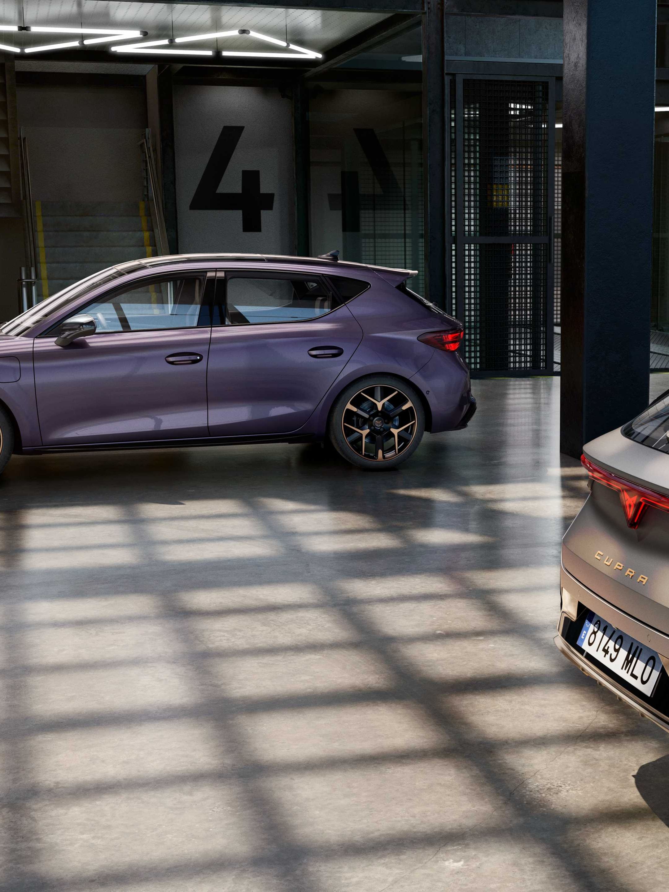 V industrijski garaži sta parkirana dva sodobna avtomobila znamke CUPRA. Eden je vijoličaste barve, drugi siv. Garaža ima velike steklene stene, skozi katere sije sončna svetloba.