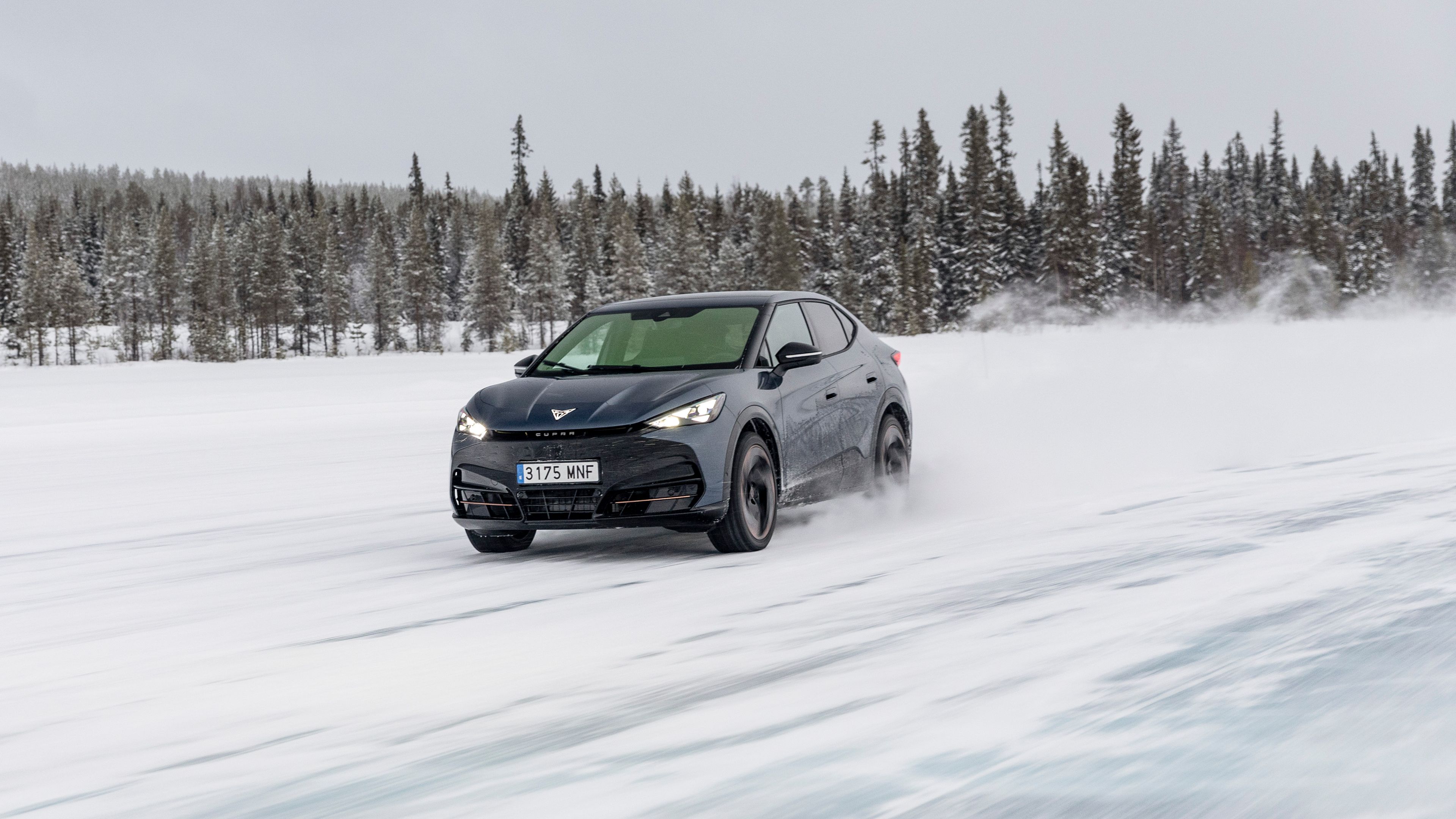 Lucas di Grassi vozi temno modri avtomobil CUPRA Tavascan po zasneženi cesti v zimskem pokrajinskem okolju. V ozadju se vidi gozd iglavcev pod oblačnim nebom. 