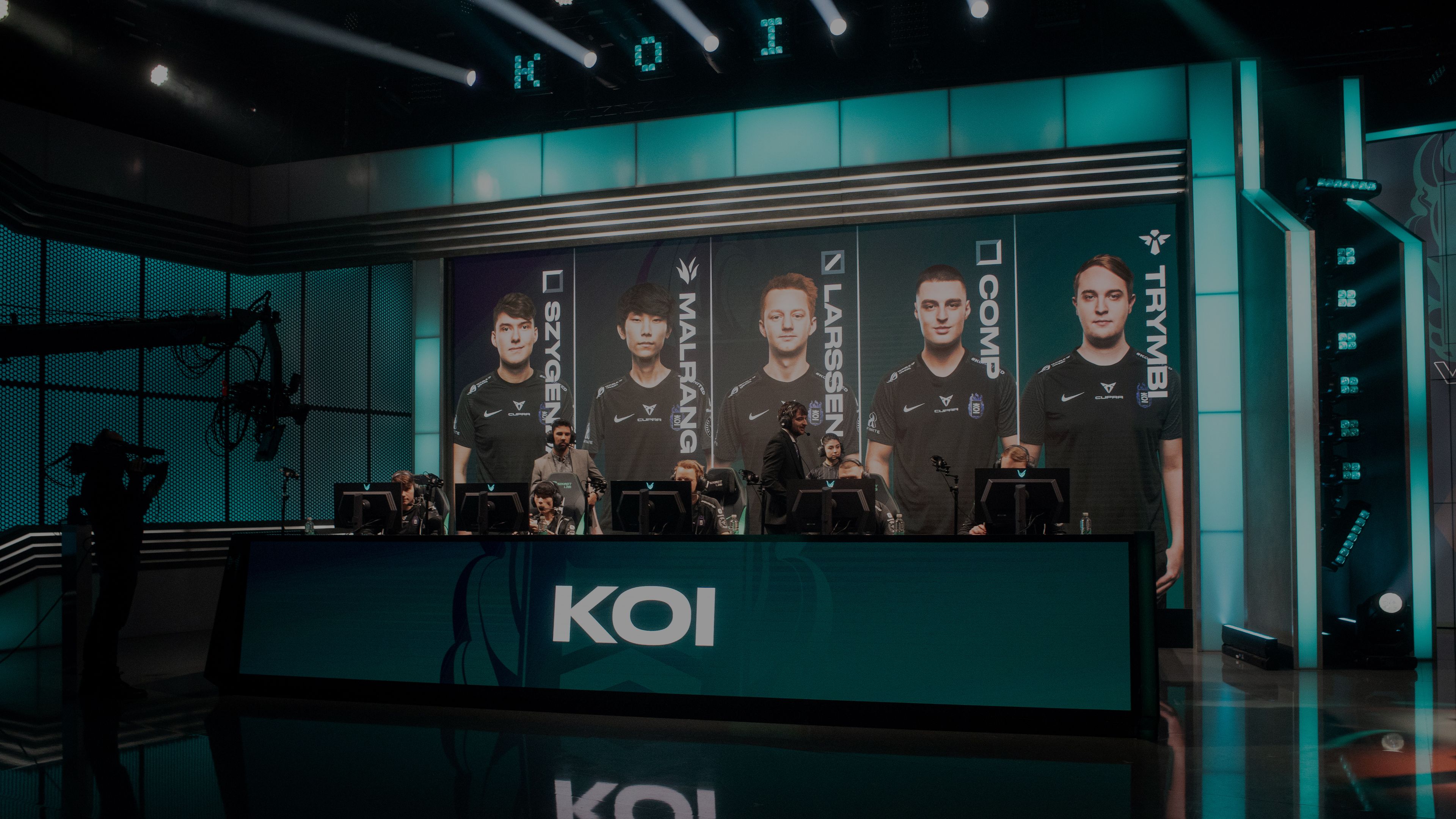 Ekipa videoigralcev sedi za računalniki na odru. Na zaslonih v ozadju so njihovi obrazi in imena. Na sprednji strani je velik napis "KOI".