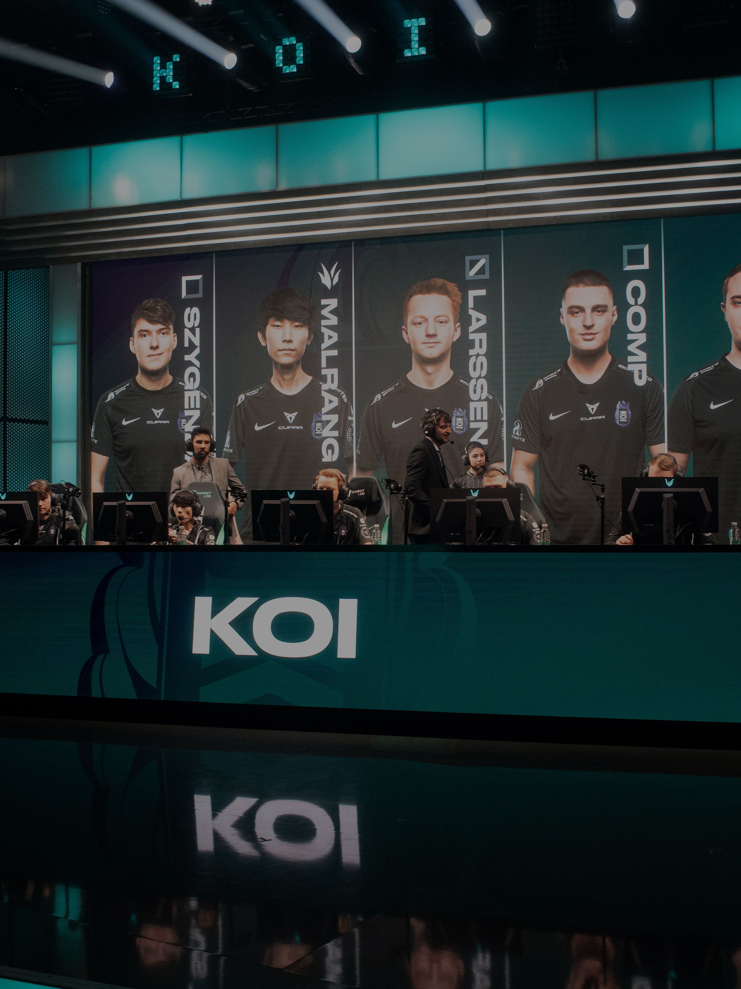 Ekipa videoigralcev sedi za računalniki na odru. Na zaslonih v ozadju so njihovi obrazi in imena. Na sprednji strani je velik napis "KOI".