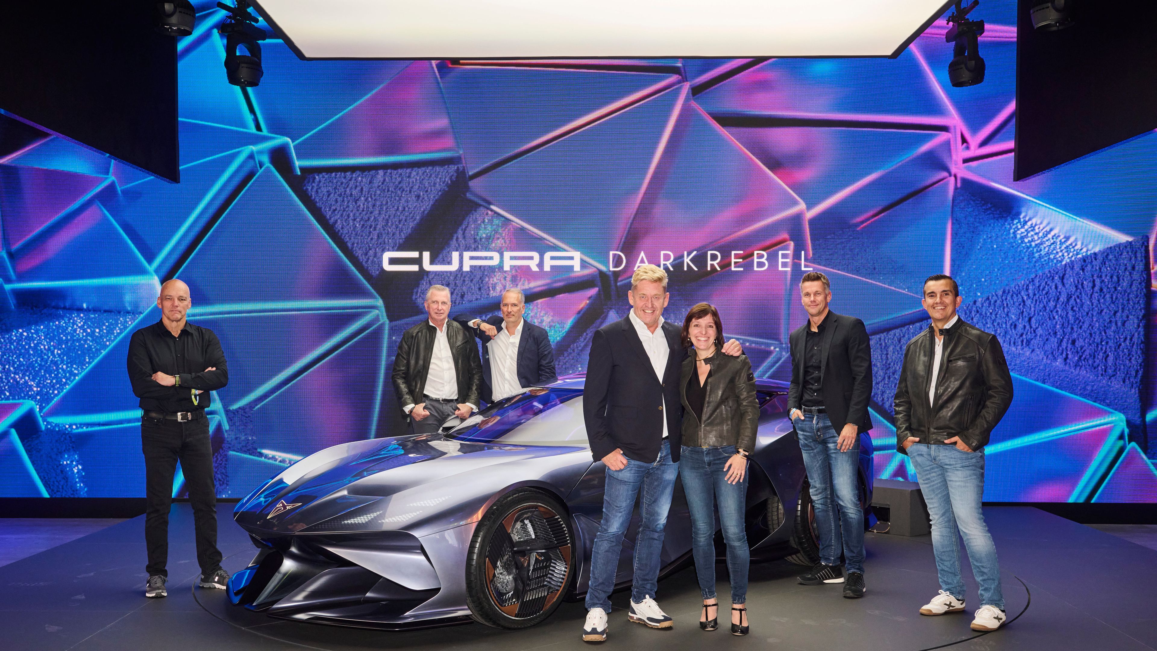 Skupina ljudi pozira pred futurističnim avtomobilom znamke CUPRA DarkRebel v sodobno osvetljenem prostoru s futurističnimi vzorci na stenah. Avtomobil je v središču pozornosti. 