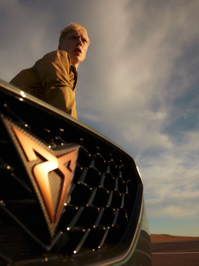 Moški stoji za avtomobilom CUPRA Formentor na katerem se jasno vidi logo znamke. Ozadje prikazuje modro-sive oblake na nebu.