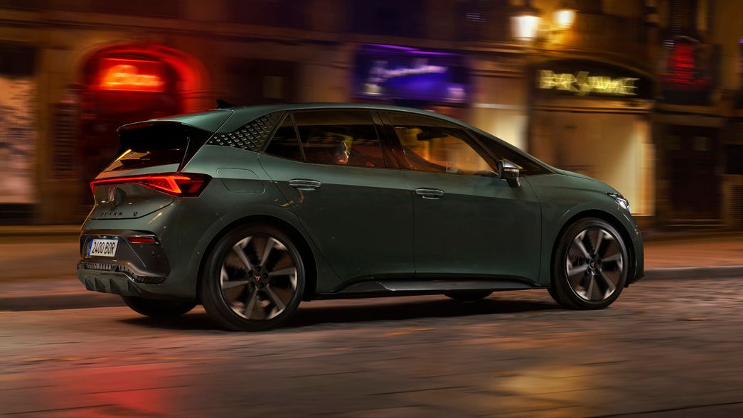 CUPRA Born VZ v temno zeleni barvi vozi po mestni ulici ponoči, osvetljeni z uličnimi lučmi. Ozadje vključuje zgradbe in trgovinske izložbe. Kolesa avtomobila so v gibanju.