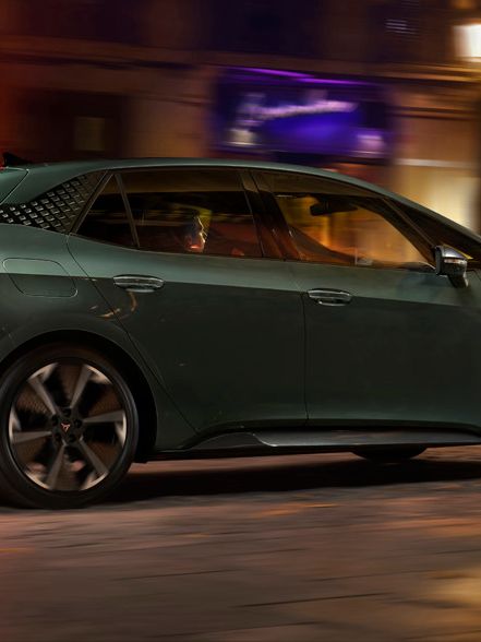 CUPRA Born VZ v temno zeleni barvi vozi po mestni ulici ponoči, osvetljeni z uličnimi lučmi. Ozadje vključuje zgradbe in trgovinske izložbe. Kolesa avtomobila so v gibanju.