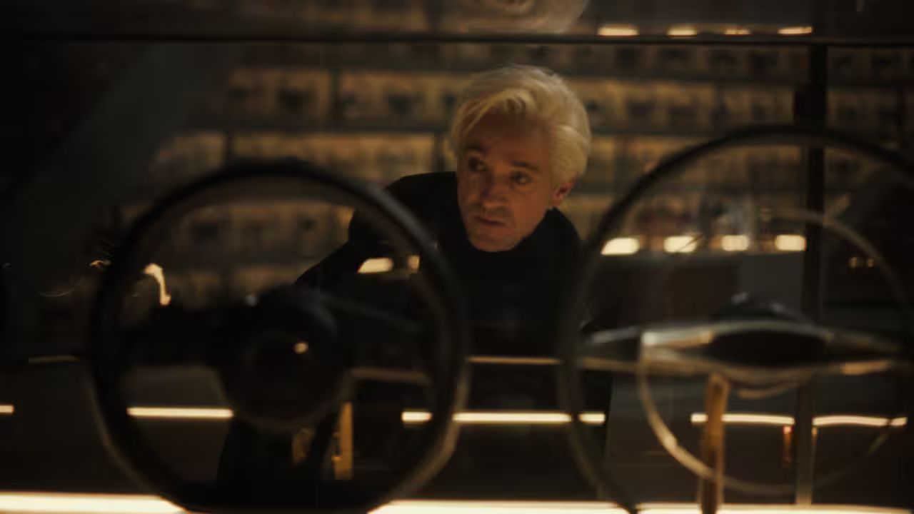Tom Felton opazuje mehanski del oziroma volan v temno osvetljenem prostoru. Prizor poudarja osredotočenost na oblikovanje in tehnične detajle.