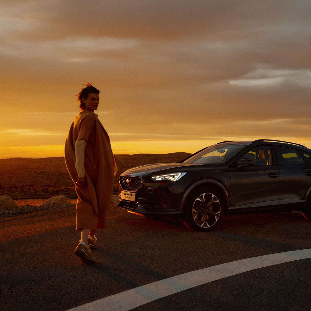 Ženska, hodi poleg parkiranega temno sivega vozila CUPRA Formentor ob sončnem zahodu. Vozilo je na cesti, v ozadju pa je vidna pokrajina.