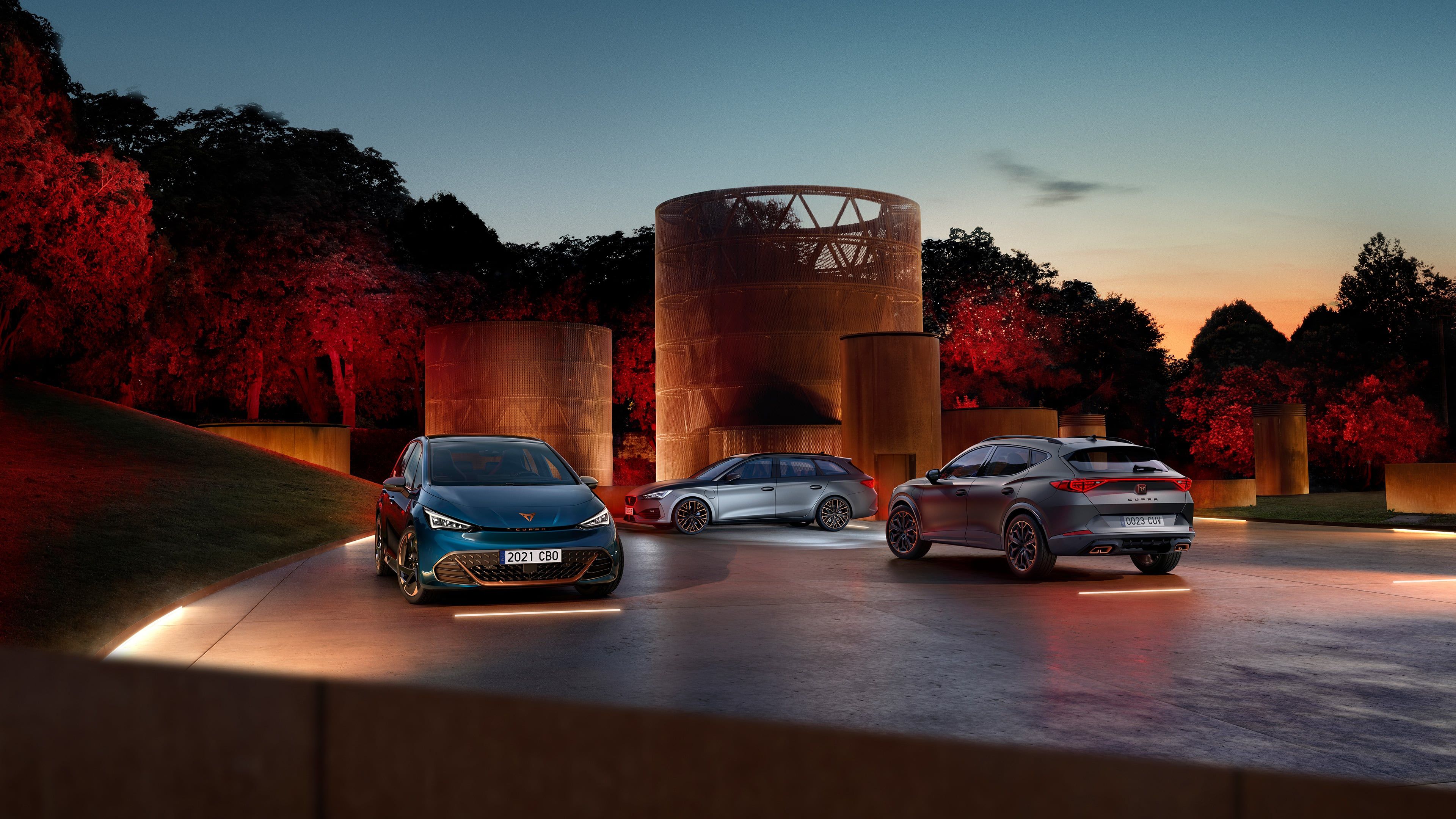 Tri sodobni avtomobili - CUPRA BORN, CUPRA LEON SP, CUPRA FORMENTOR so parkirani na dvorišču ob velikih kovinskih skulpturah v času sončnega zahoda. 