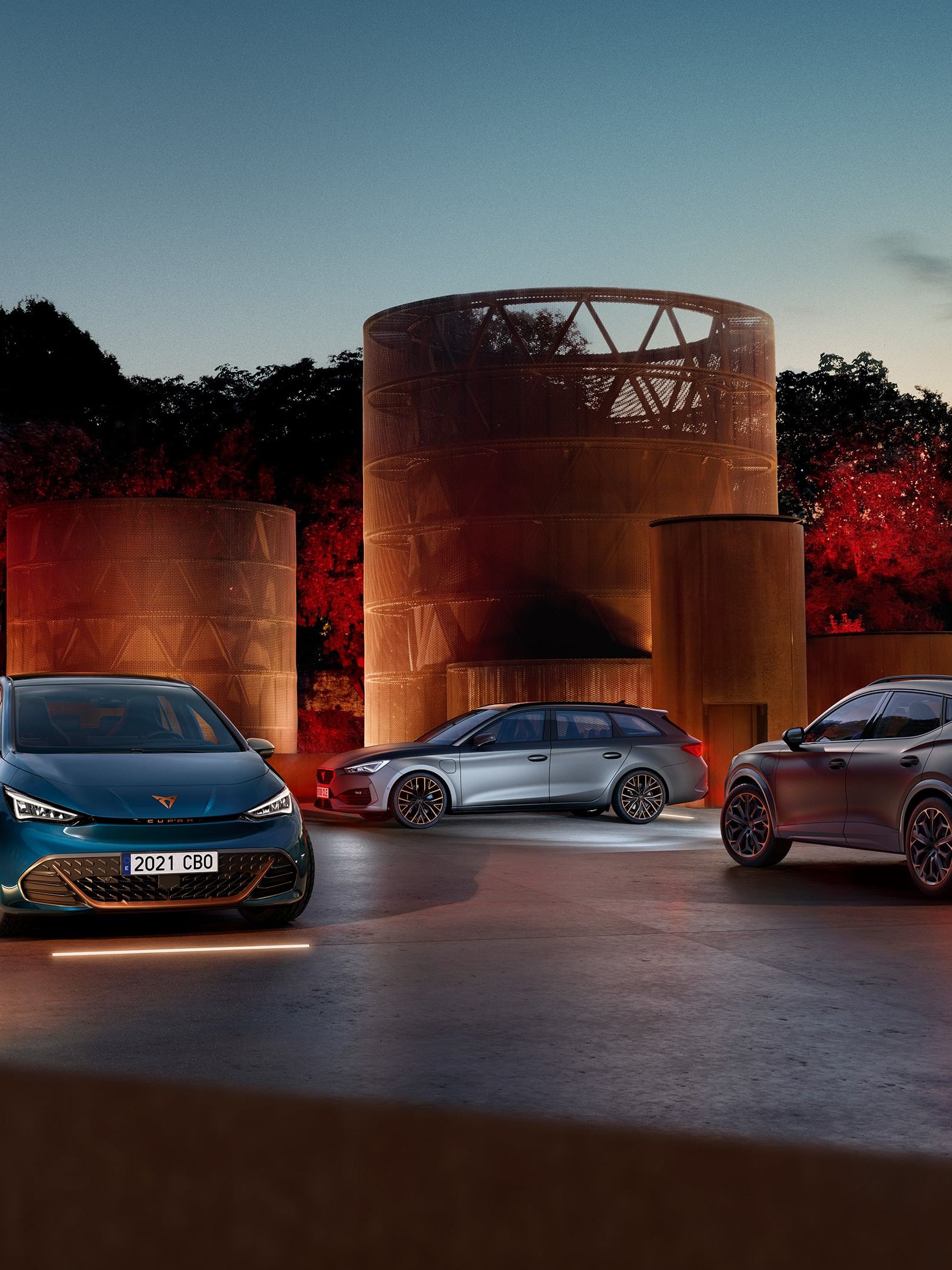 Tri sodobni avtomobili - CUPRA BORN, CUPRA LEON SP, CUPRA FORMENTOR so parkirani na dvorišču ob velikih kovinskih skulpturah v času sončnega zahoda. 
