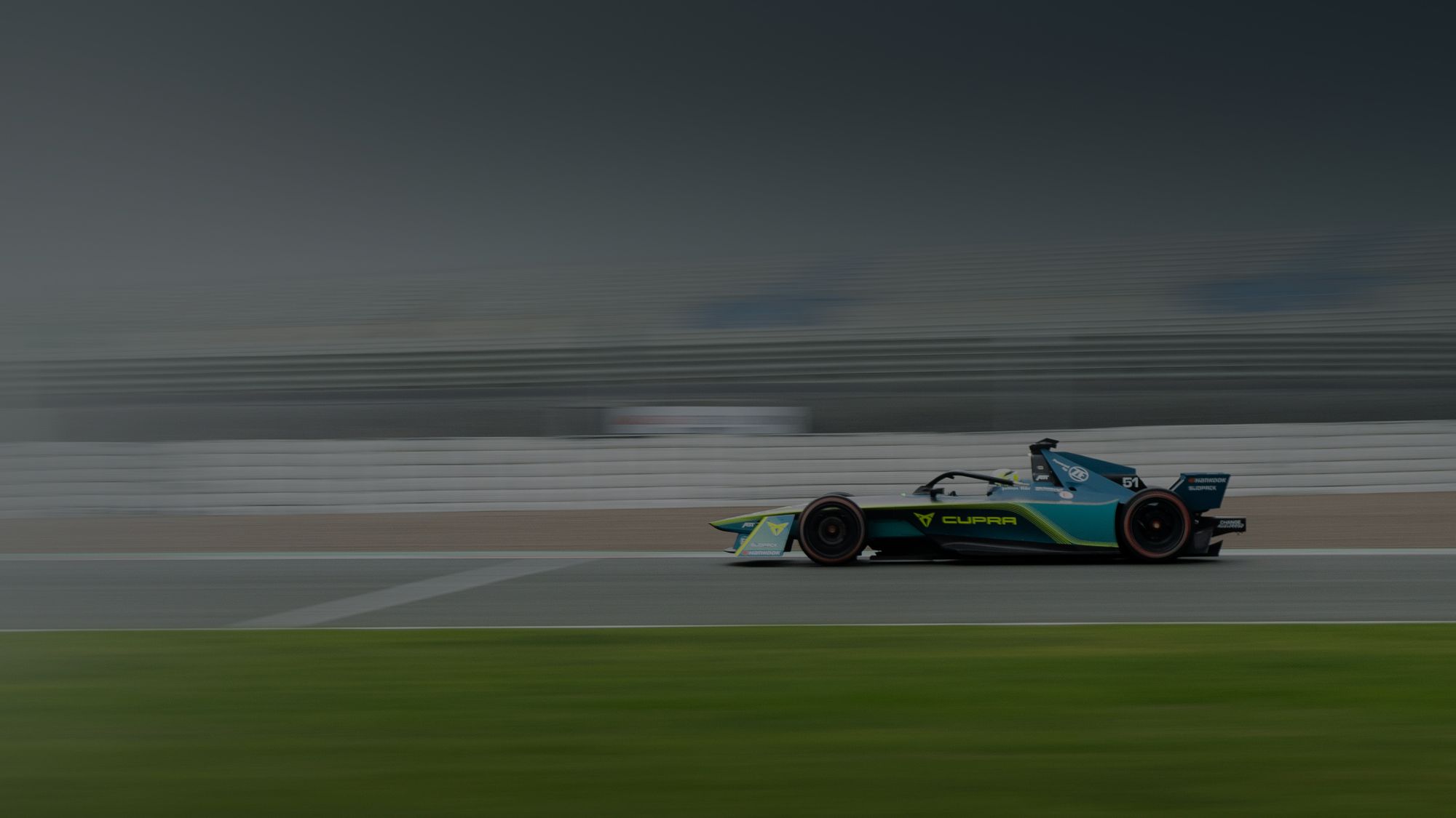 Dirkalnik formule 1 v hitrem gibanju na dirkalni stezi. Ozadje je zamegljeno zaradi hitrosti. Spredaj je vidna zelena trava ob stezi.