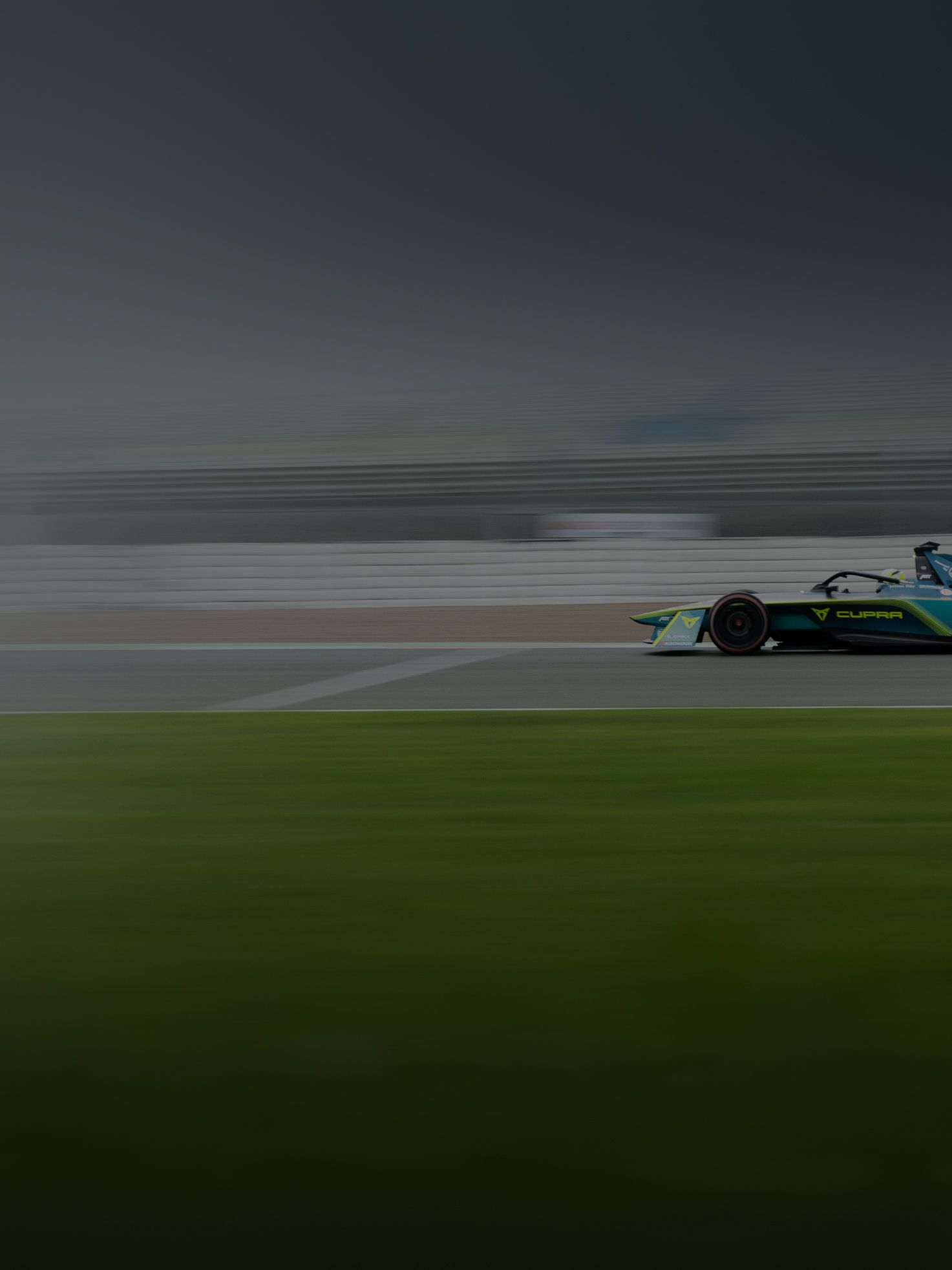 Dirkalnik formule 1 v hitrem gibanju na dirkalni stezi. Ozadje je zamegljeno zaradi hitrosti. Spredaj je vidna zelena trava ob stezi.