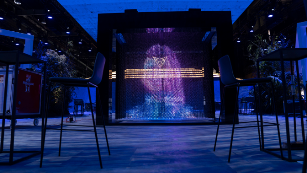 Interaktivna inštalacija prikazuje barvni svetlobni prikaz na panelu iz več luči. Namestitev je obdana s stoli in mizami v modernem prostoru z modro osvetlitvijo. 