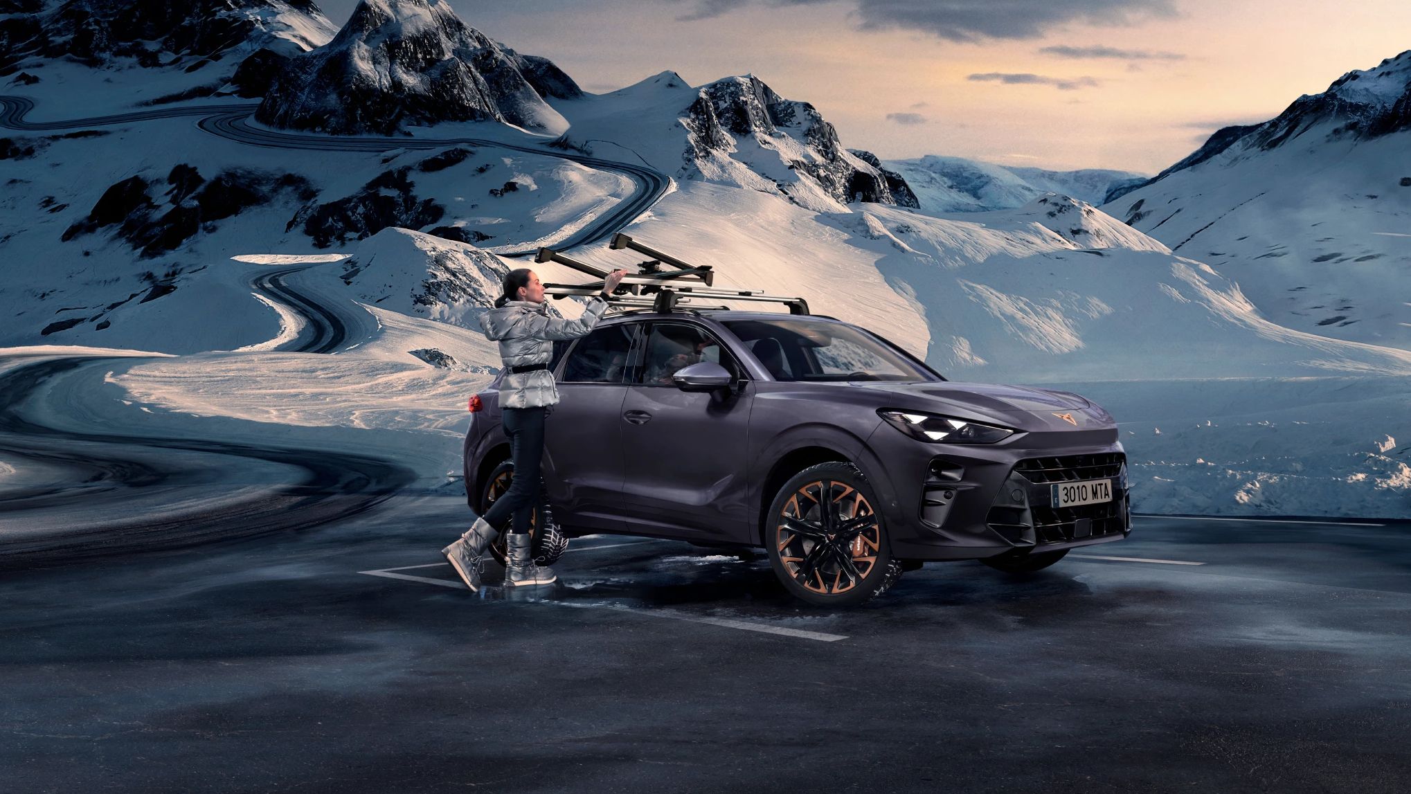 CUPRA Terramar stoji na zasneženem gorskem območju ob vijugasti cesti. Ženska namešča smuči na strešne nosilce vozila. V ozadju so visoki zasneženi vrhovi pod oblačnim nebom.