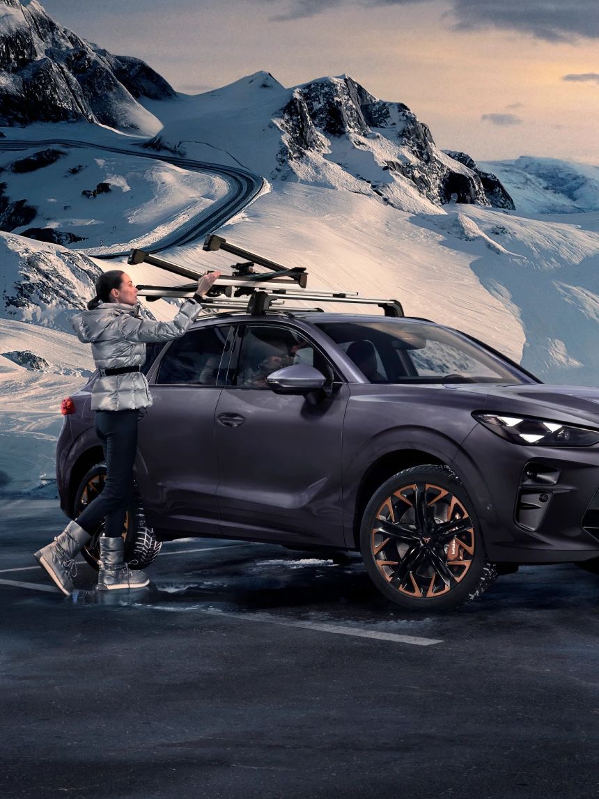 CUPRA Terramar stoji na zasneženem gorskem območju ob vijugasti cesti. Ženska namešča smuči na strešne nosilce vozila. V ozadju so visoki zasneženi vrhovi pod oblačnim nebom.