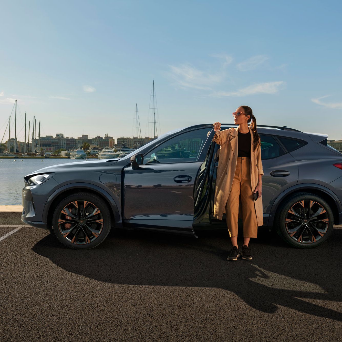 Ženska stoji ob odprtih vratih sivega vozila CUPRA Formentor PA, parkiranega ob obali. V ozadju je marinina pokrajina z jadralnimi čolni na vodi in poslopji pod svetlim nebom.