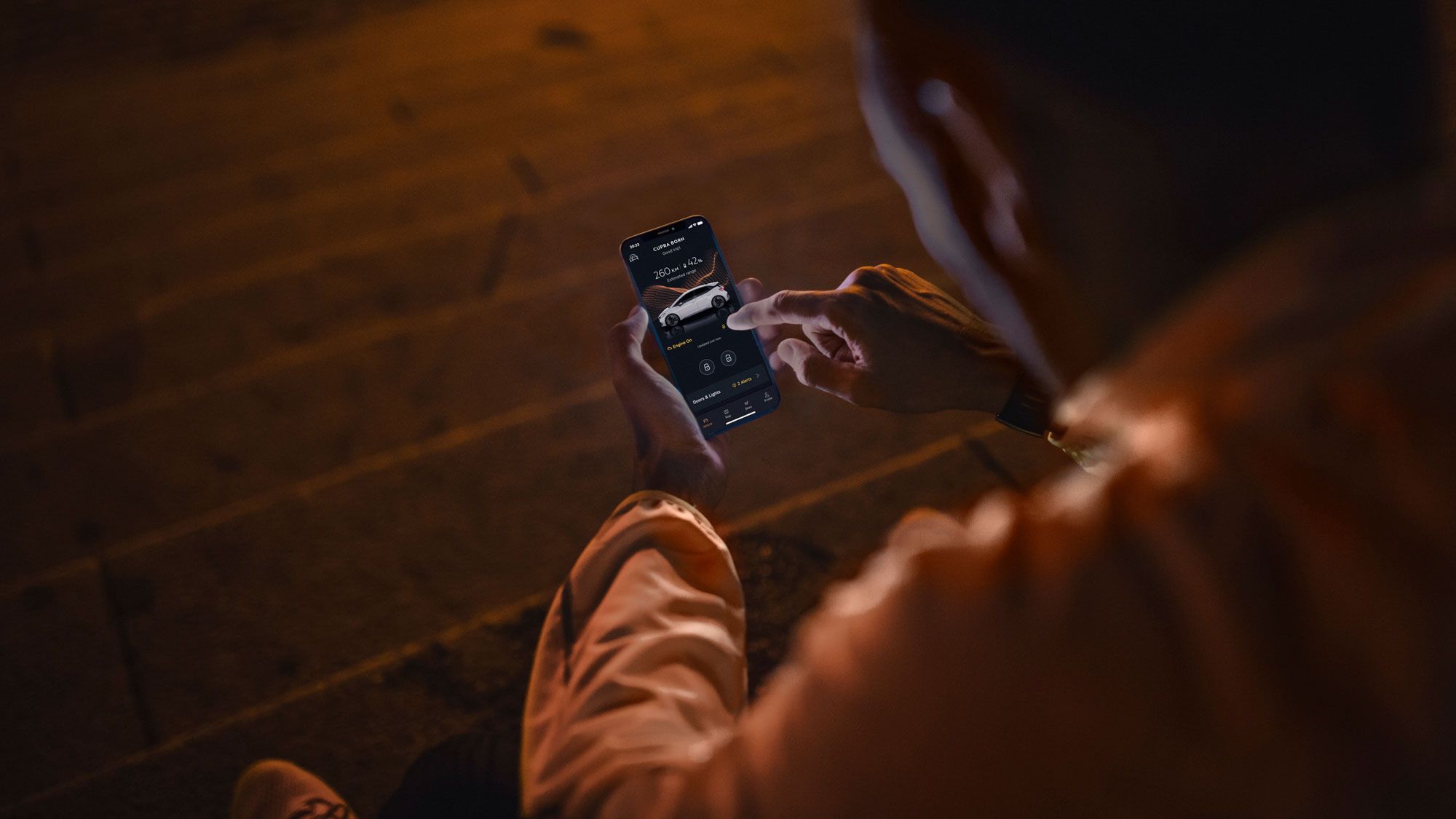 Oseba sedi na stopnicah v temi in na svojem pametnem telefonu uporablja aplikacijo, aplikacijo CUPRA za model Born. Njegov prst je usmerjen na zaslon, kjer se vidi slika avtomobila.