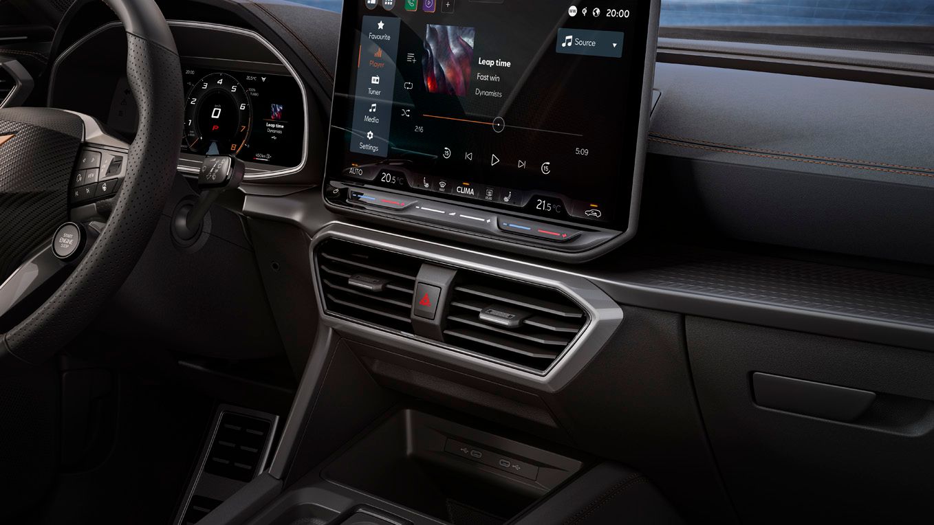 Notranjost avtomobila CUPRA Formentor PA PHEV VZ, z modernim zaslonom na dotik na armaturni plošči, na katerem je prikazana navigacija. V ozadju se vidi mestno obzorje ob vodi med sončnim zahodom.