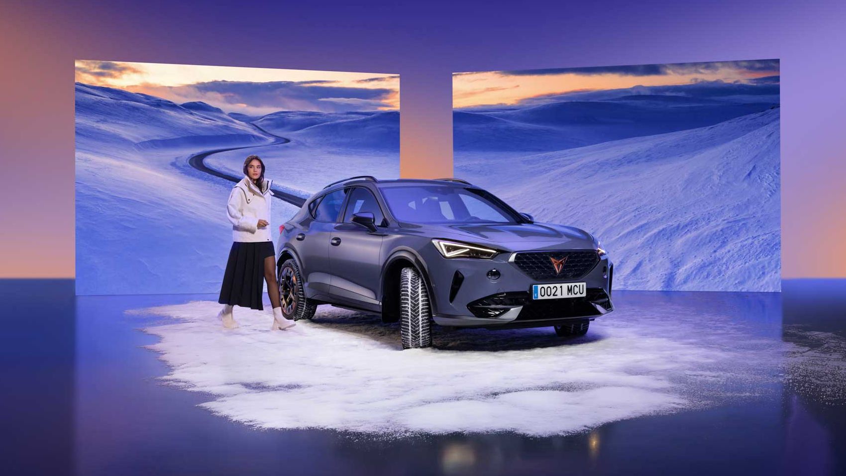 V ospredju je črn CUPRA Formentor postavljen na simuliran sneg, obkrožen s panoramskimi zasloni, ki prikazujejo zasneženo pokrajino. Oseba v belem oblačilu stoji ob avtomobilu in gleda proti zaslonom. Ozadje je obarvano z modro-oranžnimi barvnimi prehodi.