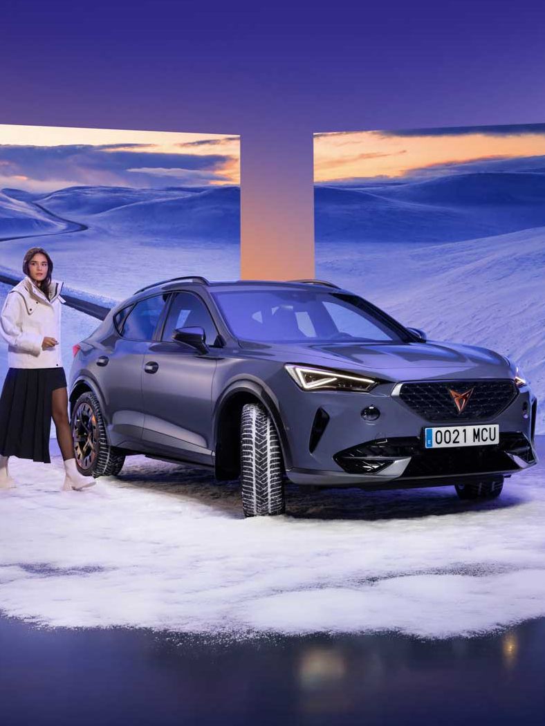 V ospredju je črn CUPRA Formentor postavljen na simuliran sneg, obkrožen s panoramskimi zasloni, ki prikazujejo zasneženo pokrajino. Oseba v belem oblačilu stoji ob avtomobilu in gleda proti zaslonom. Ozadje je obarvano z modro-oranžnimi barvnimi prehodi.