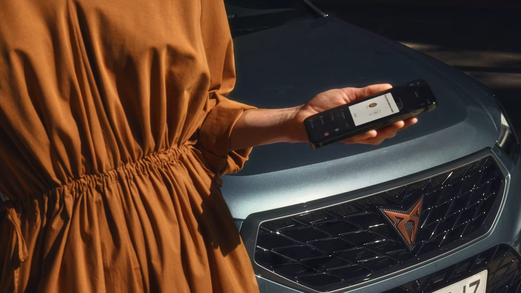 Oseba v oker obleki stoji ob avtomobilu CUPRA Ateca in drži telefon z aplikacijo "My CUPRA app" na zaslonu. Avtomobil je sive barve. 