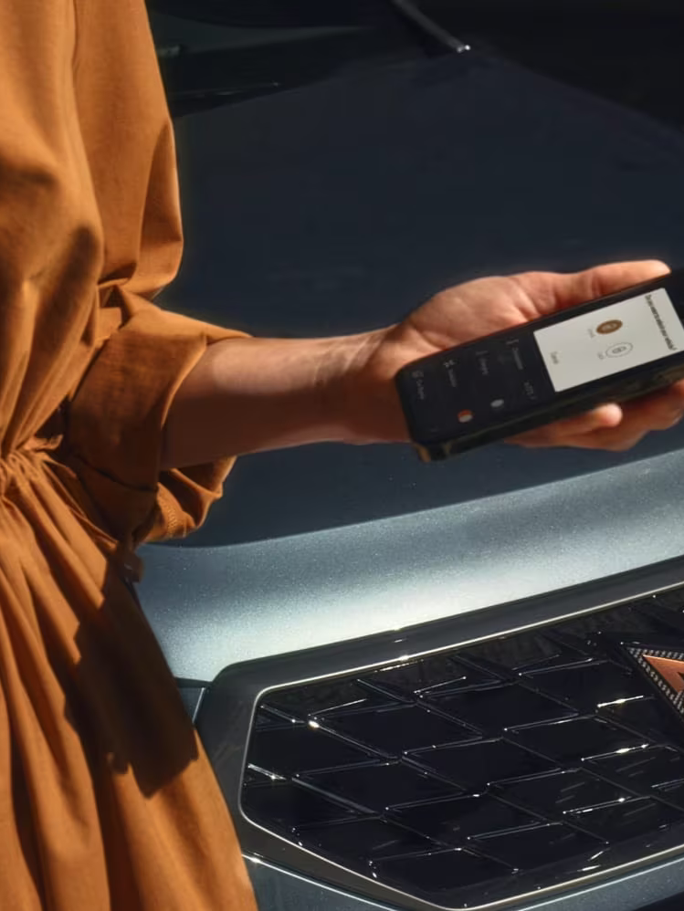 Oseba v oker obleki stoji ob avtomobilu CUPRA Ateca in drži telefon z aplikacijo "My CUPRA app" na zaslonu. Avtomobil je sive barve. 