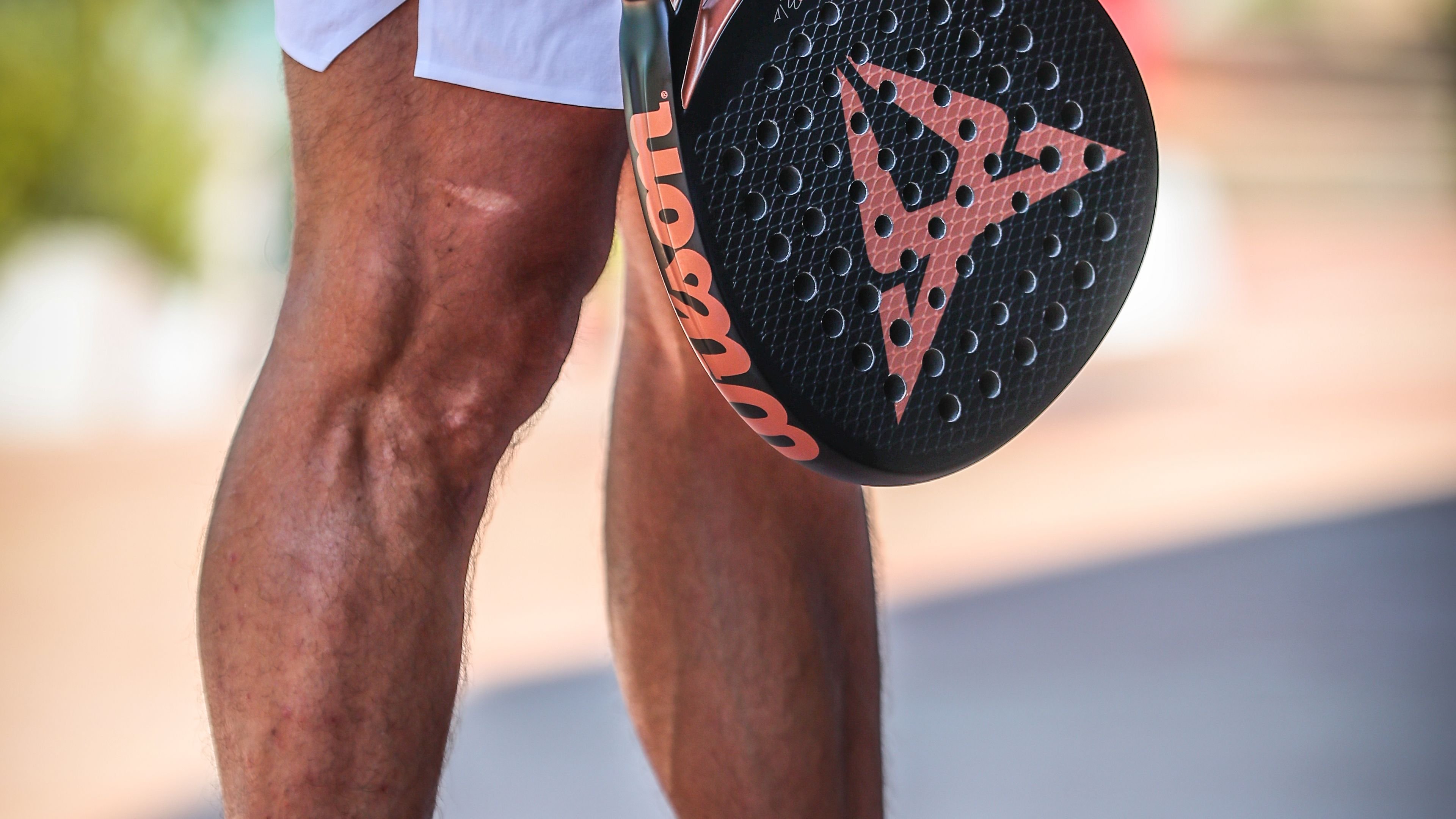 Moški stoji s padel loparjem v roki z logotipom znamke CUPRA, oblečen v belo teniško opremo in rdeče športne copate z logotipom znamke Wilson.