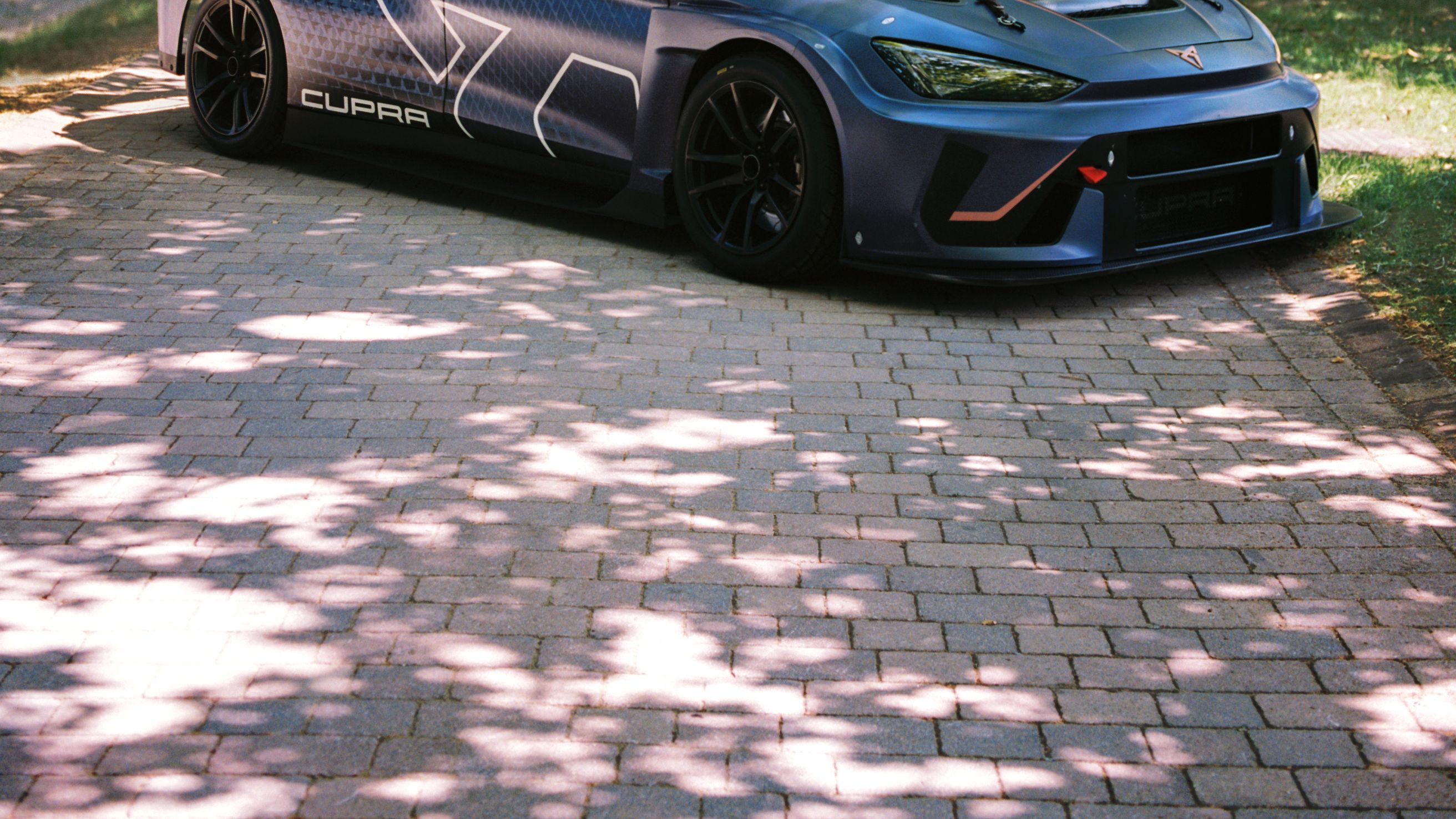 Oseba v jahalni opremi z oranžno čelado in očali sedi na pokrovu modrega športnega avtomobila ter drži vajeti, kot da jezdi avto, med vožnjo po drevoredni cesti.