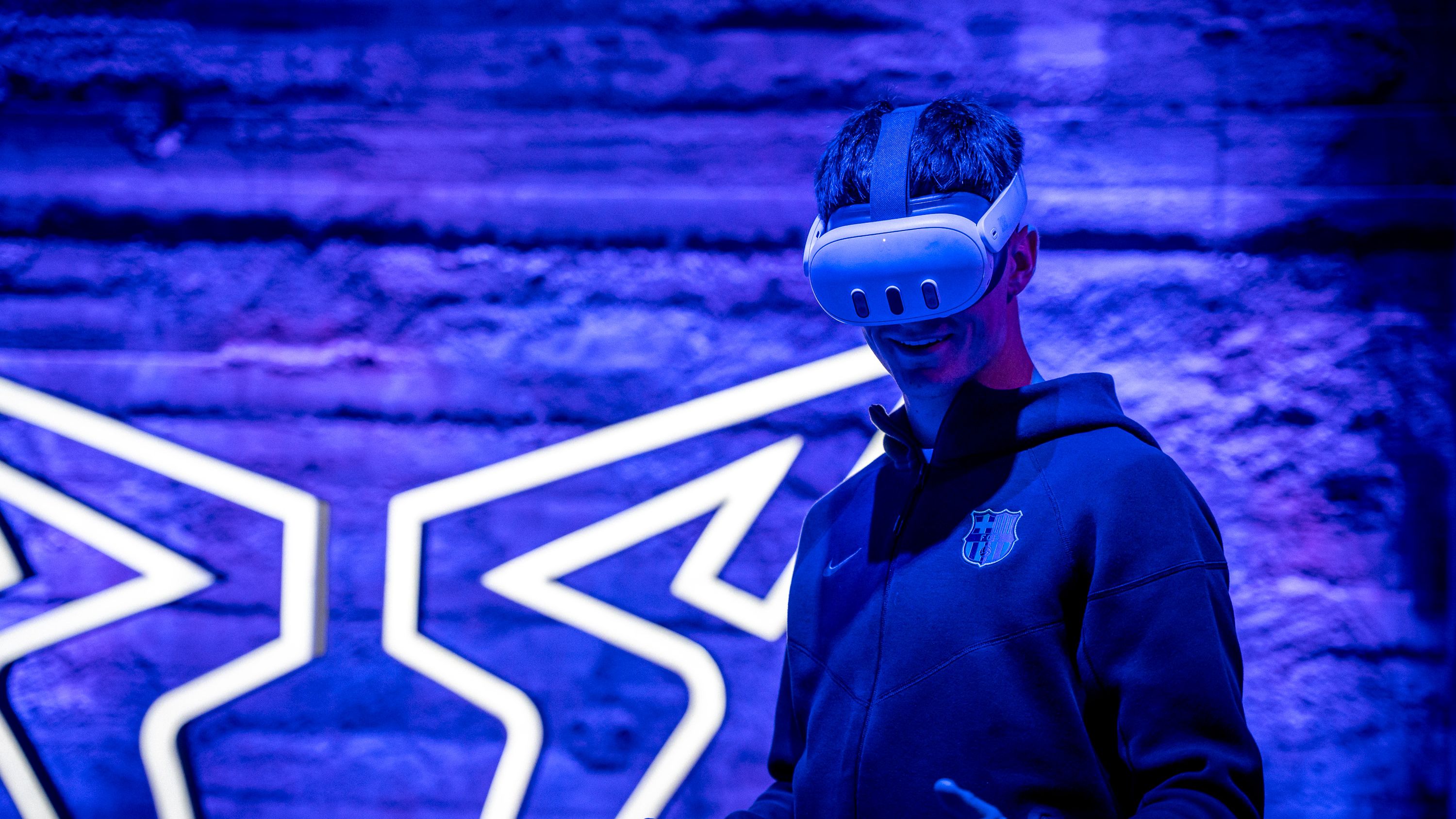 Igralec kluba Barcelona nosi VR očala v temnem prostoru z modro-vijolično osvetlitvijo. Za njo je osvetljen logotip znamke CUPRA na steni.