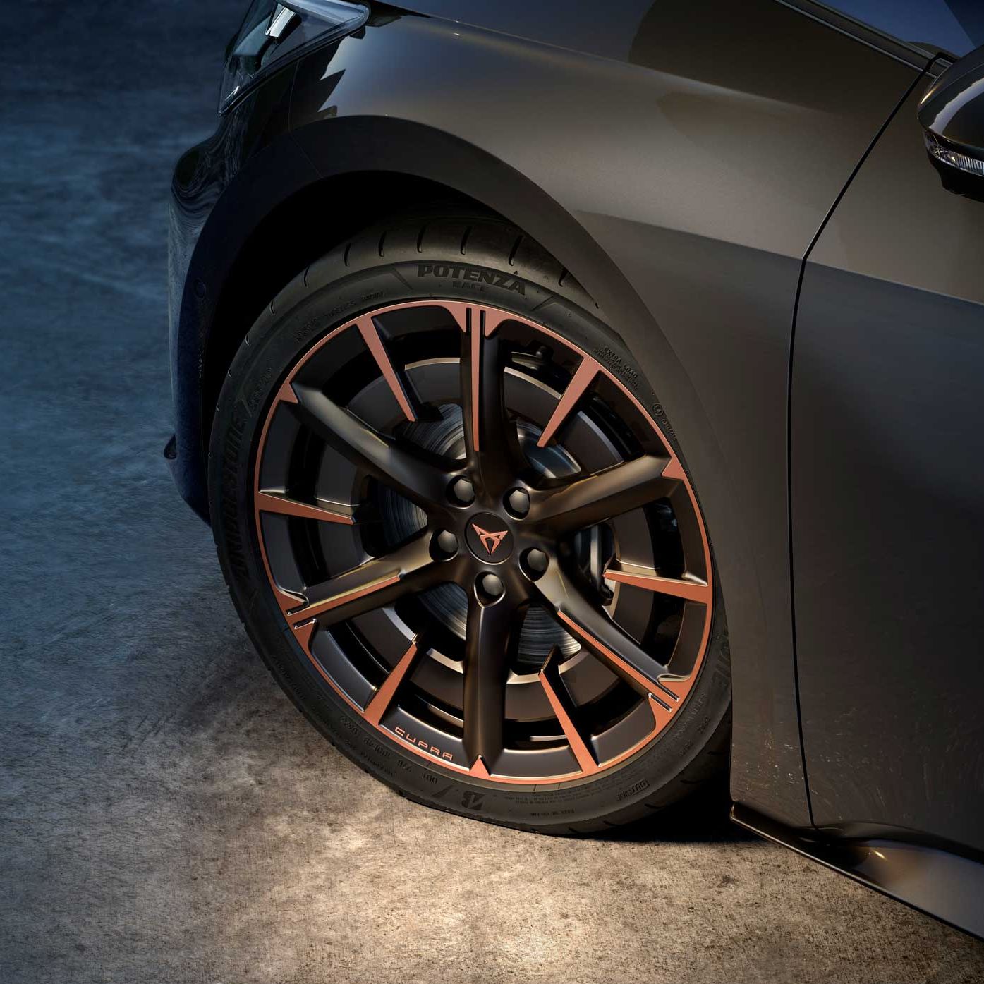 Detaljen sprednjega kolesa CUPRA Born z velikimi športnimi platišči v mat črni barvi z bakrenimi poudarki. Avto je temno sive barve, osvetljen od spodaj, kar poudari obliko platišča in pnevmatike. 