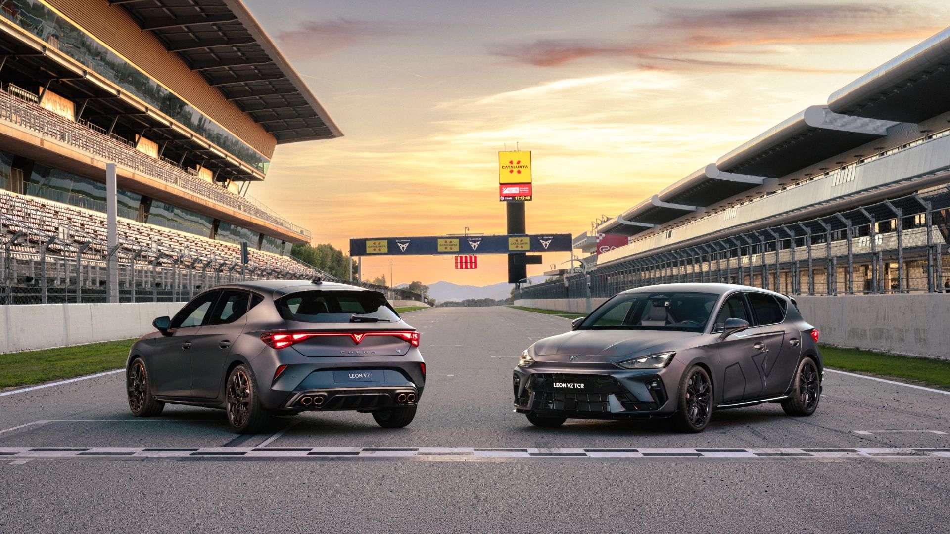 Na sliki sta prikazana dva športna avtomobila CUPRA Leon, parkirana na progi dirkališča. V ozadju je tribuna z sedišči, jasen sončni zahod pa osvetljuje prizorišče. 