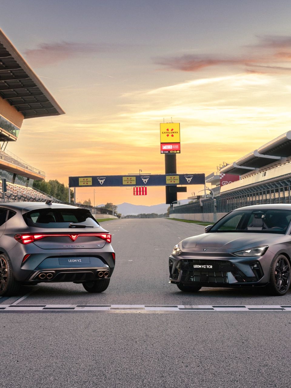 Na sliki sta prikazana dva športna avtomobila CUPRA Leon, parkirana na progi dirkališča. V ozadju je tribuna z sedišči, jasen sončni zahod pa osvetljuje prizorišče. 