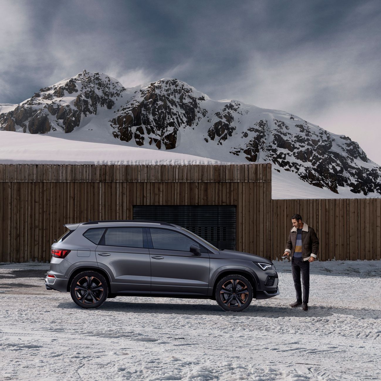 Moški stoji poleg sivega avtomobila CUPRA Ateca, na zasneženem parkirišču, zadaj pa je lesena stavba in snežni gorovje.