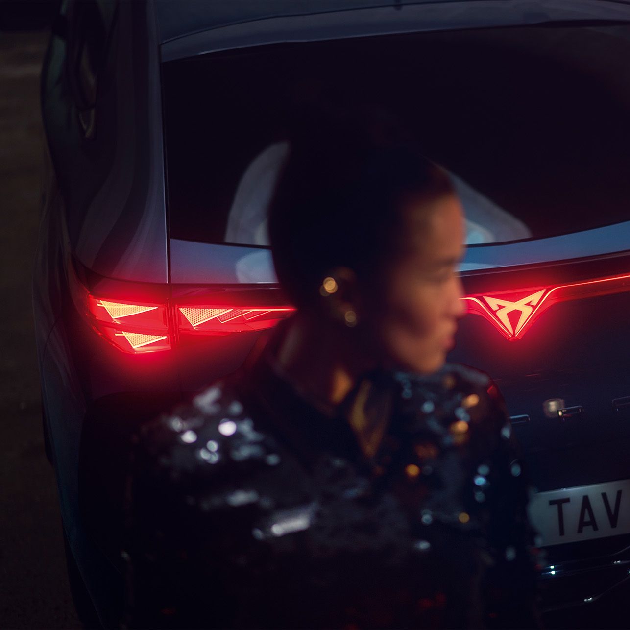 Ženska stoji pred električnim Cupra Tavascan avtomobilom z vklopljenimi zadnjimi lučmi, ki so v obliki črke Y. Na registrski tablici avtomobila je napis "TAV".