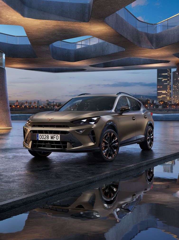 Parkiran CUPRA Formentor PA na zunanjem sodobnem parkirišču z zanimivo arhitekturo. V ozadju se vidi mestna panorama ob reki. Nebo je modro ob sončnem zahodu.