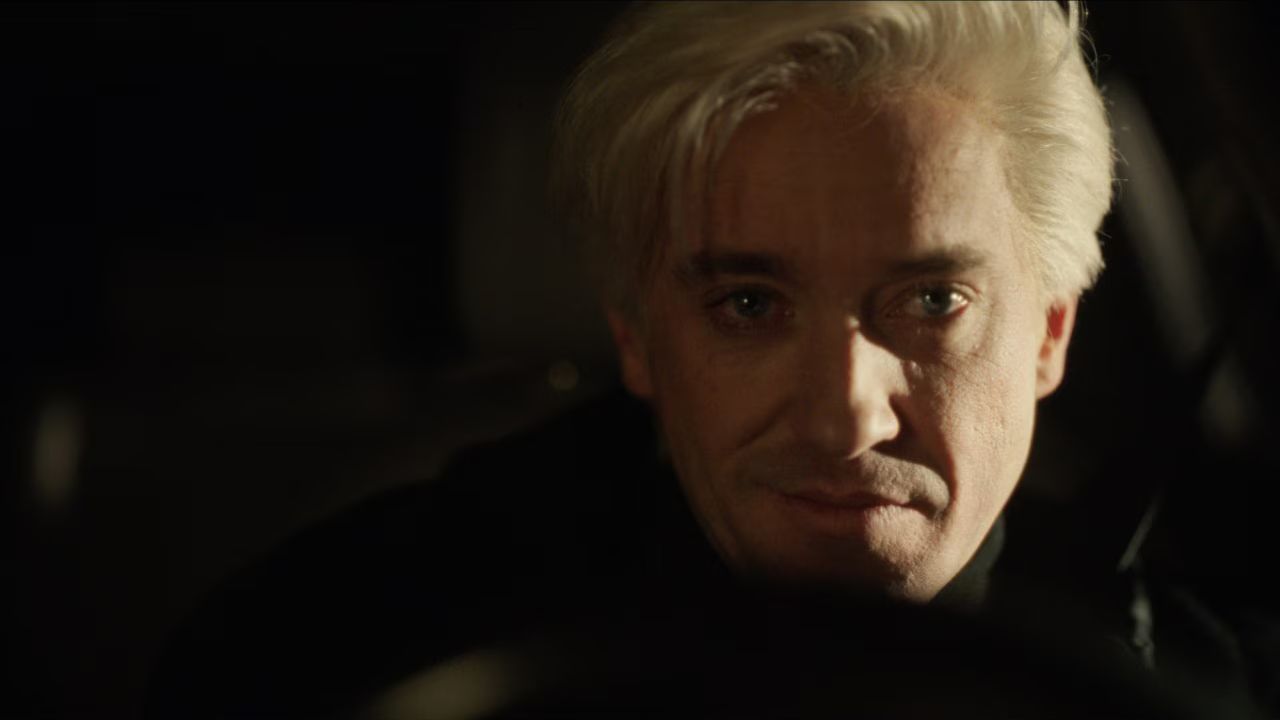 Tom Felton sedi v zatemnjenem prostoru vozila in gleda proti kameri. Mehka svetloba poudarja dramatično in skrivnostno vzdušje prizora.