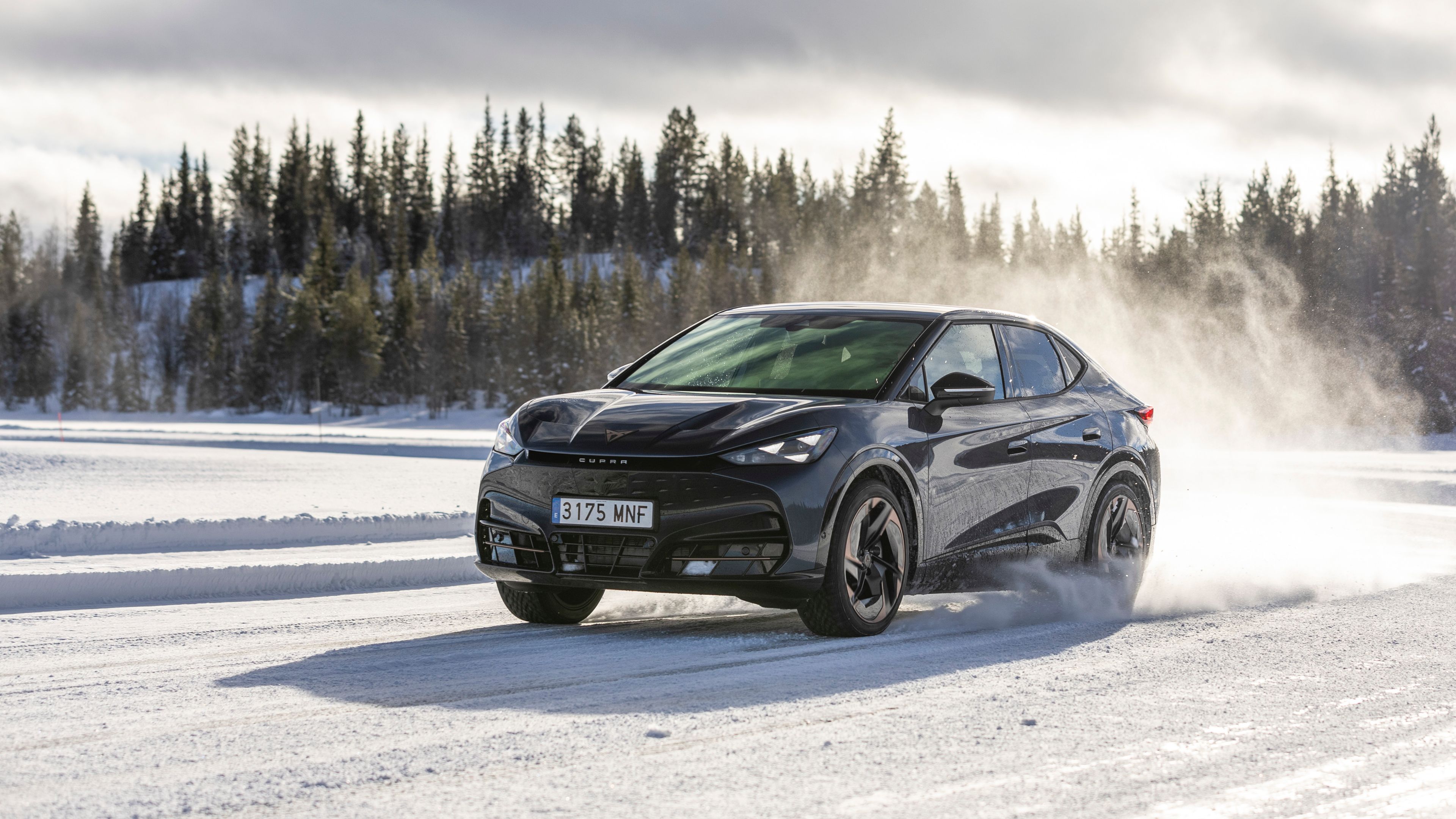 Črn avtomobil CUPRA vozi po zasneženi cesti v zimskem pokrajinskem okolju. V ozadju se vidi gozd iglavcev pod oblačnim nebom. 