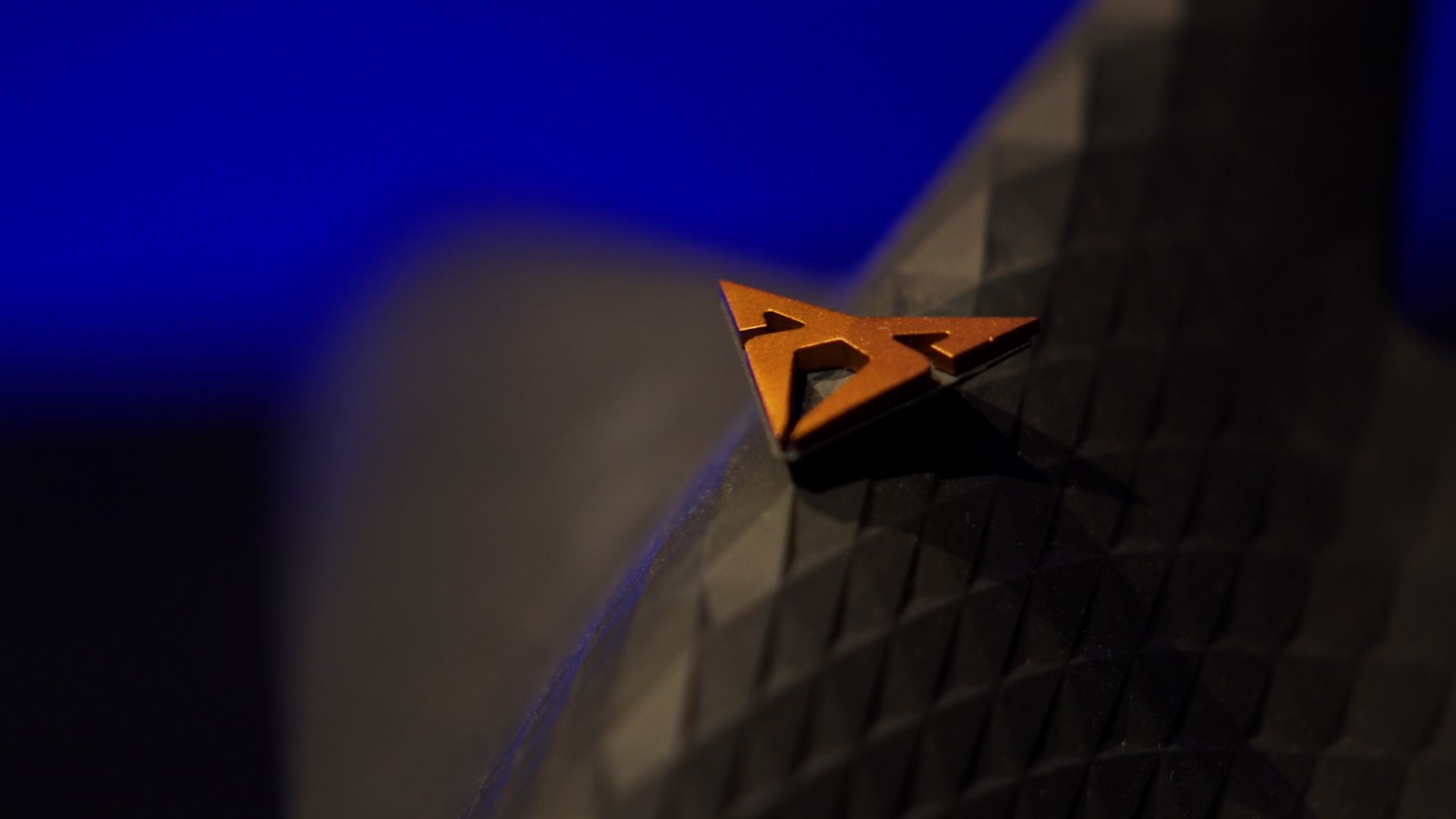 Prikaz logotipa znamke CUPRA, ki leži na črni teksturirani površini s temno modrim ozadjem.