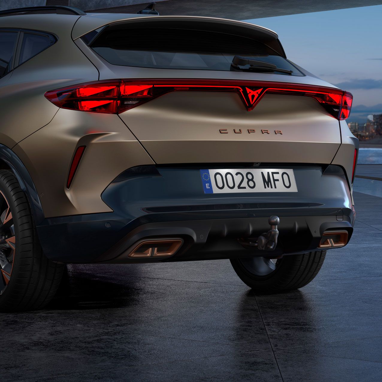 Zadnji del vozila CUPRA Formentor PA je parkiran v mestu ob reki, z osvetljenimi stavbami v ozadju. Vozilo je v mat kovinsko zlati barvi.