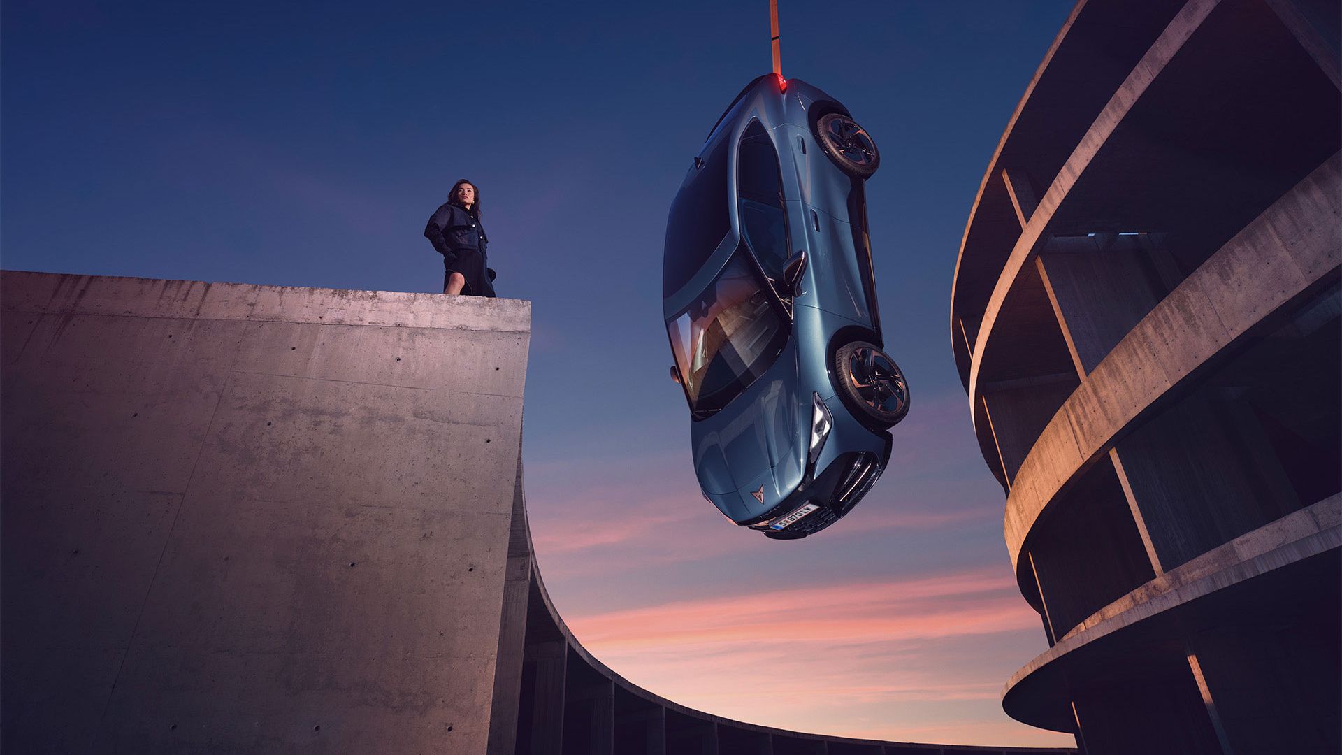 Modri CUPRA Tavascan visi v zraku, pritrjen na žerjav. Ob njem stoji ženska na robu betonske konstrukcije, v ozadju pa je zaobljena večnadstropna parkirna hiša v mraku.