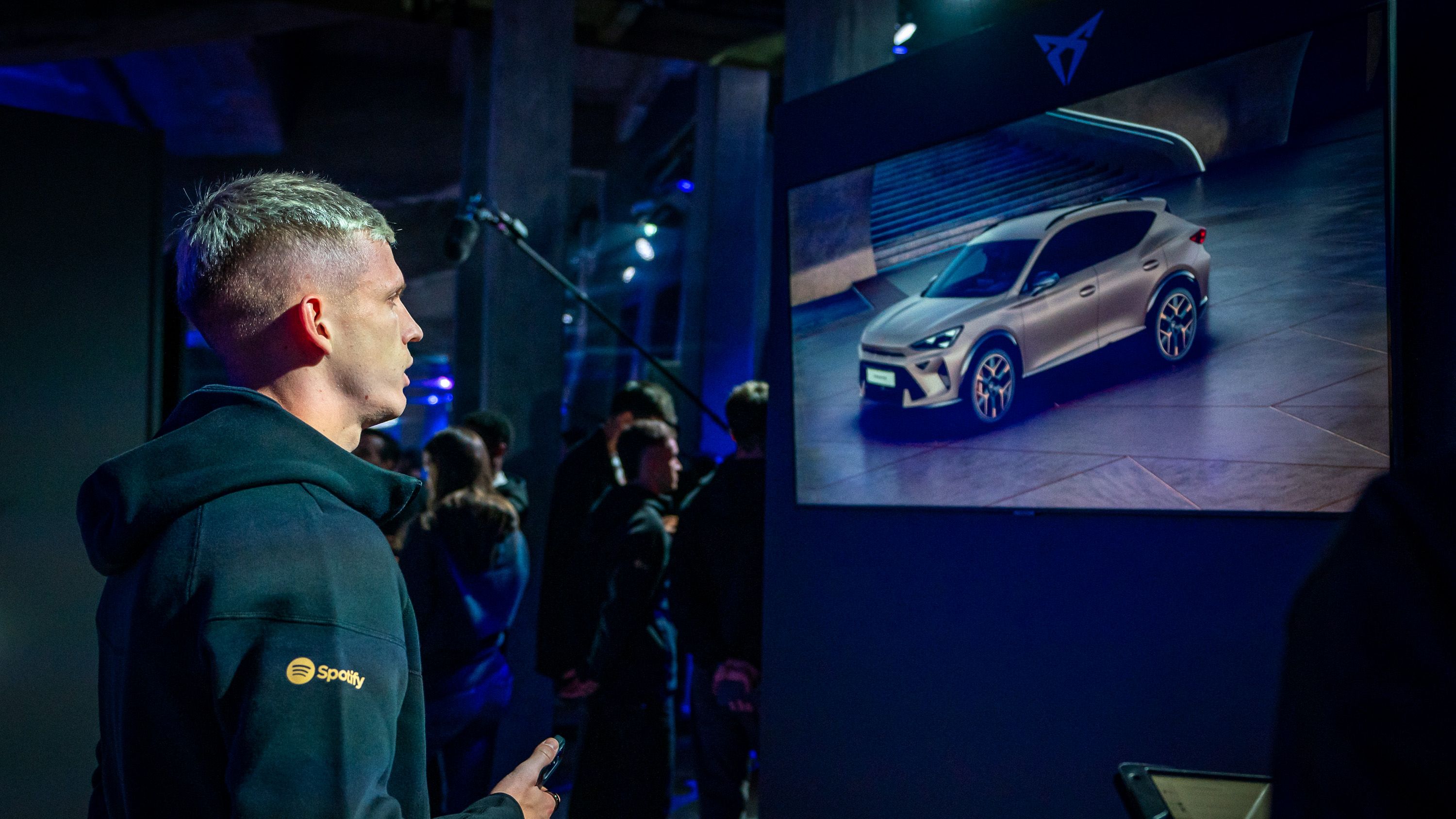 Igralec FC Barcelona stoji pred zaslonom, na katerem je prikazan avtomobil CUPRA Terramar. V ozadju se zadržuje več ljudi v temno osvetljenem prostoru.