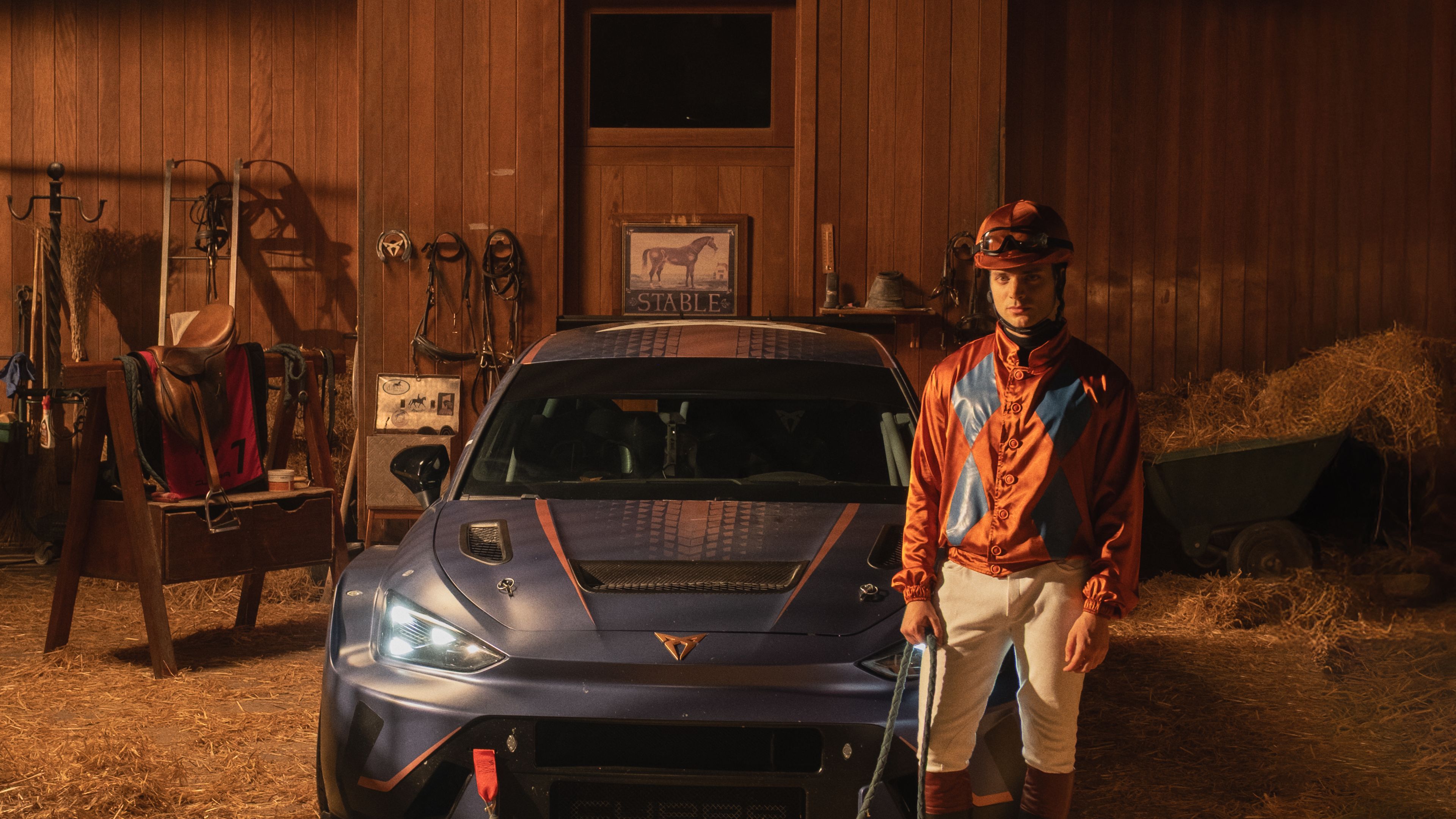 Oseba stoji ob dirkalnem avtomobilu znamke CUPRA v prostoru, ki spominja na hlev. Oblečena je v jahalno opremo, v ozadju pa so vidni balini sena in orodje za konje.