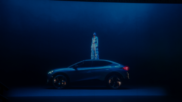 Oseba v svetlih oblačilih stoji na strehi temnega avtomobila CUPRA v prostoru z modro osvetlitvijo. Avto ima športni dizajn in velike platišča. Ozadje je enobarvno in temno.