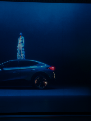 Oseba v svetlih oblačilih stoji na strehi temnega avtomobila CUPRA v prostoru z modro osvetlitvijo. Avto ima športni dizajn in velike platišča. Ozadje je enobarvno in temno.