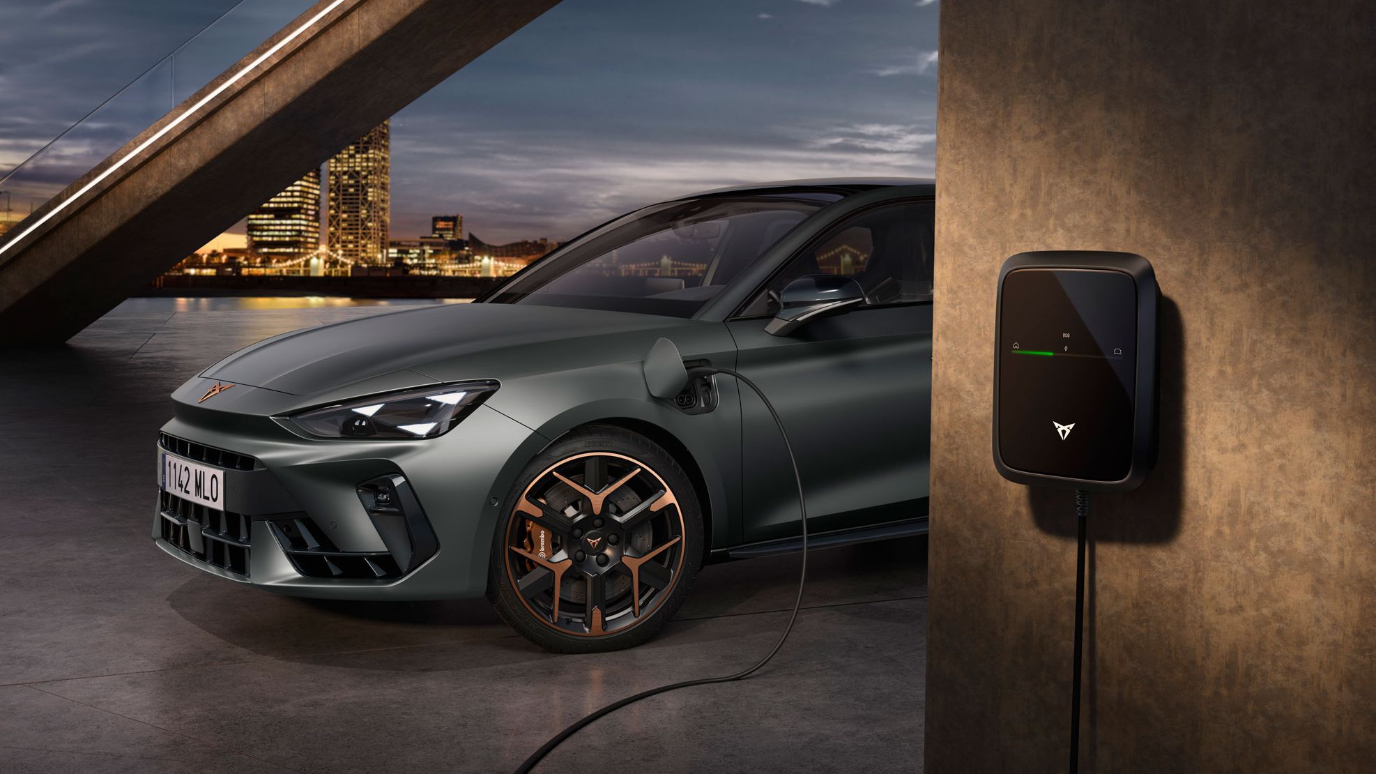 CUPRA Leon PA je priključen na polnilno postajo. V ozadju se vidi mestna pokrajina v večernem času. Vozilo je parkirano na asfaltni površini.