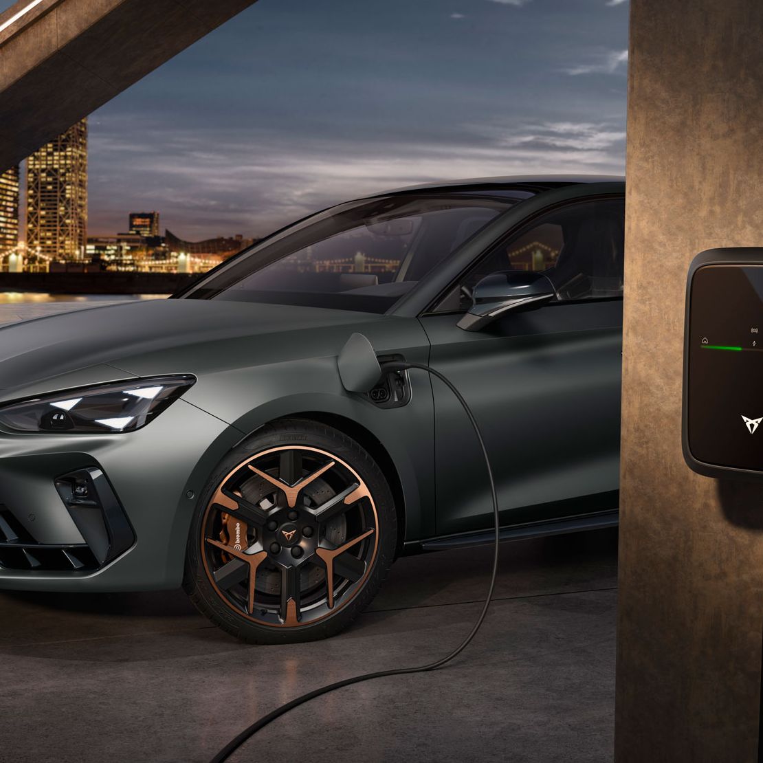 CUPRA Leon PA je priključen na polnilno postajo. V ozadju se vidi mestna pokrajina v večernem času. Vozilo je parkirano na asfaltni površini.