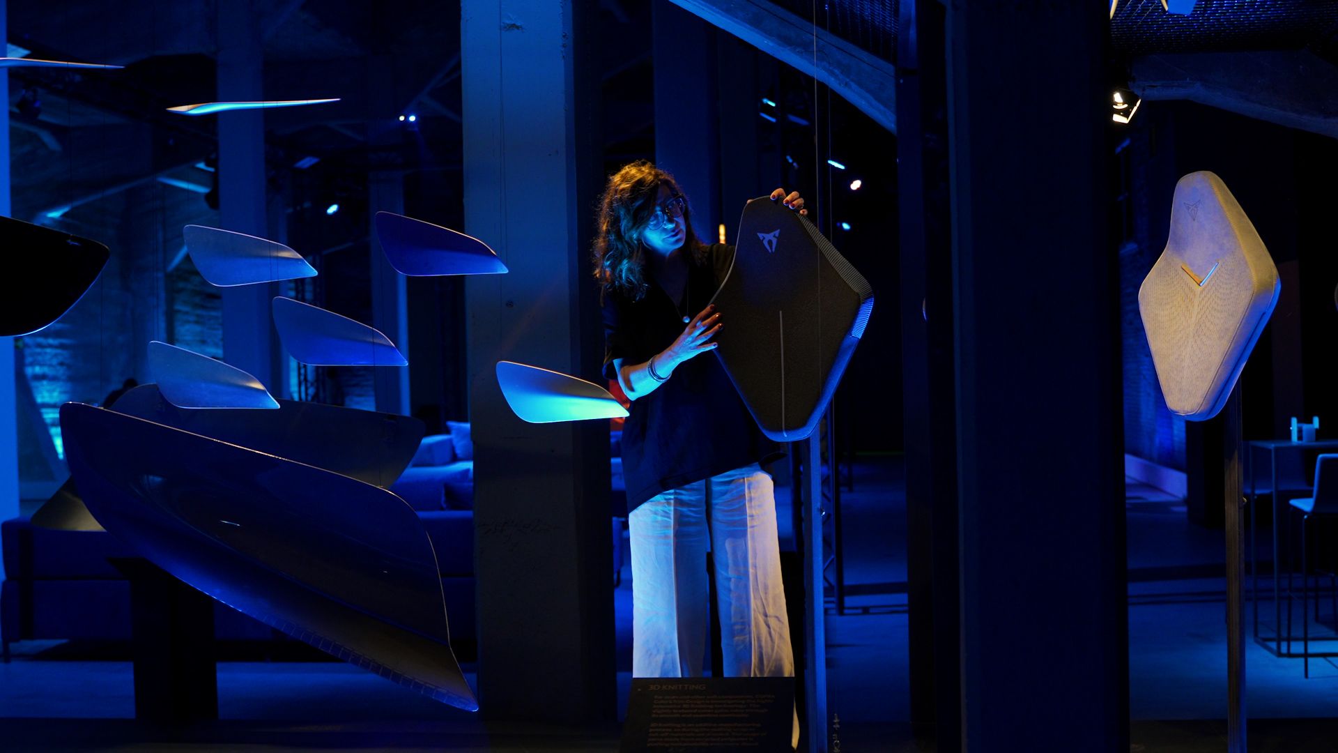 Oseba v modro osvetljenem prostoru interaktivno sodeluje z umetniško inštalacijo z visečimi avtomobilskimi sedeži. Na sedežu je logotip znamke CUPRA.