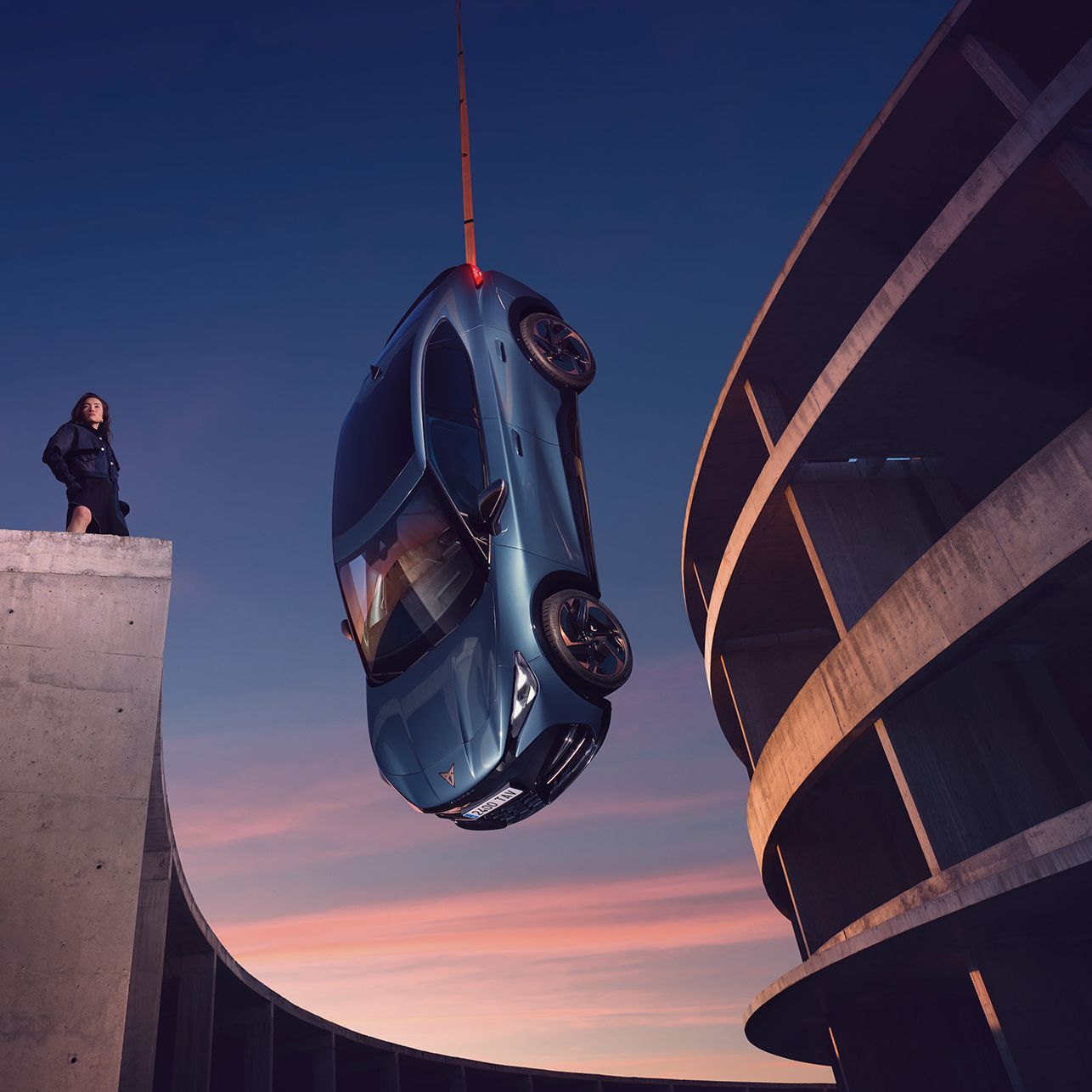 Ženska stoji na robu betonske zgradbe in opazuje modri CUPRA Tavascan VZ, ki visi v zraku, obešen na žerjav. V ozadju se vidi sončni zahod.