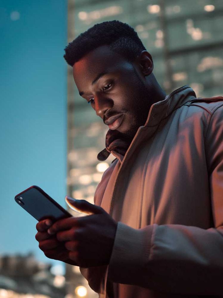 Moški stoji na mestni ulici ponoči in drži pametni telefon. V ozadju so osvetljene steklene stavbe. Svetloba z ulice in stavb ustvarja zanimiv kontrast.