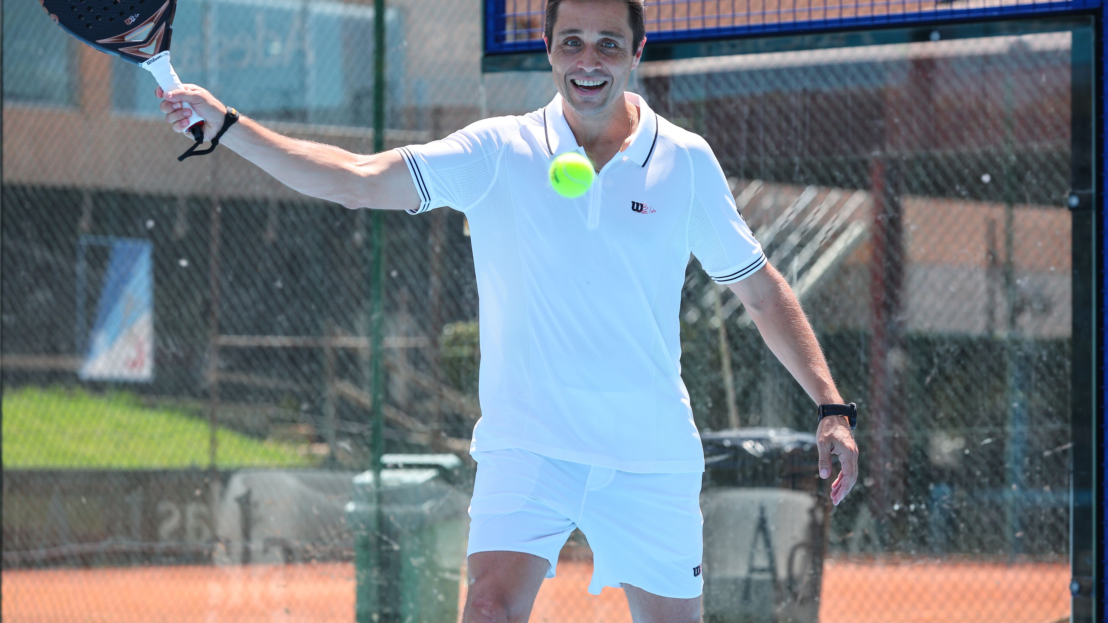 Fernando Belasteguín v beli športni opremi igra padel. Ima dvignjeno roko in udarja žogico s padel loparjem. V ozadju je mreža in igrišče za padel. Vide se logotipi CUPRA, Wilson in Bela.