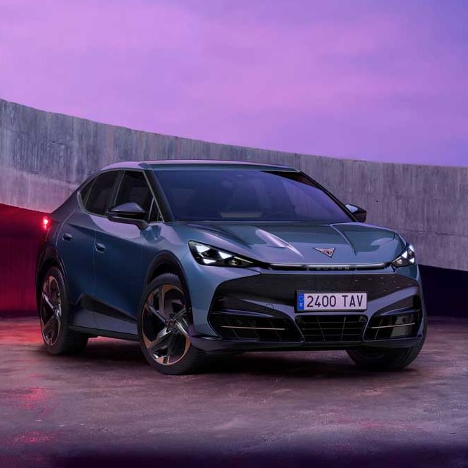 Temen Cupra Tavascan stoji na odprtem prostoru, obdanim z betonskim zidom, ob sončnem zahodu. Avto je športen, temno sive barve, z modernim dizajnom. Nebo je obarvano v vijoličnih in rožnatih odtenkih.