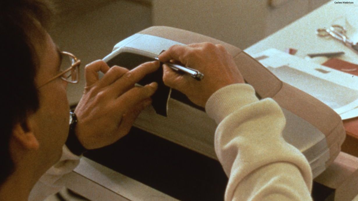 Oseba s kiparskim orodjem oblikuje majhen model avtomobila znamke SEAT. Model je na mizi, v ozadju pa so različni dokumenti in orodja. Slika je stara približno 40 let.